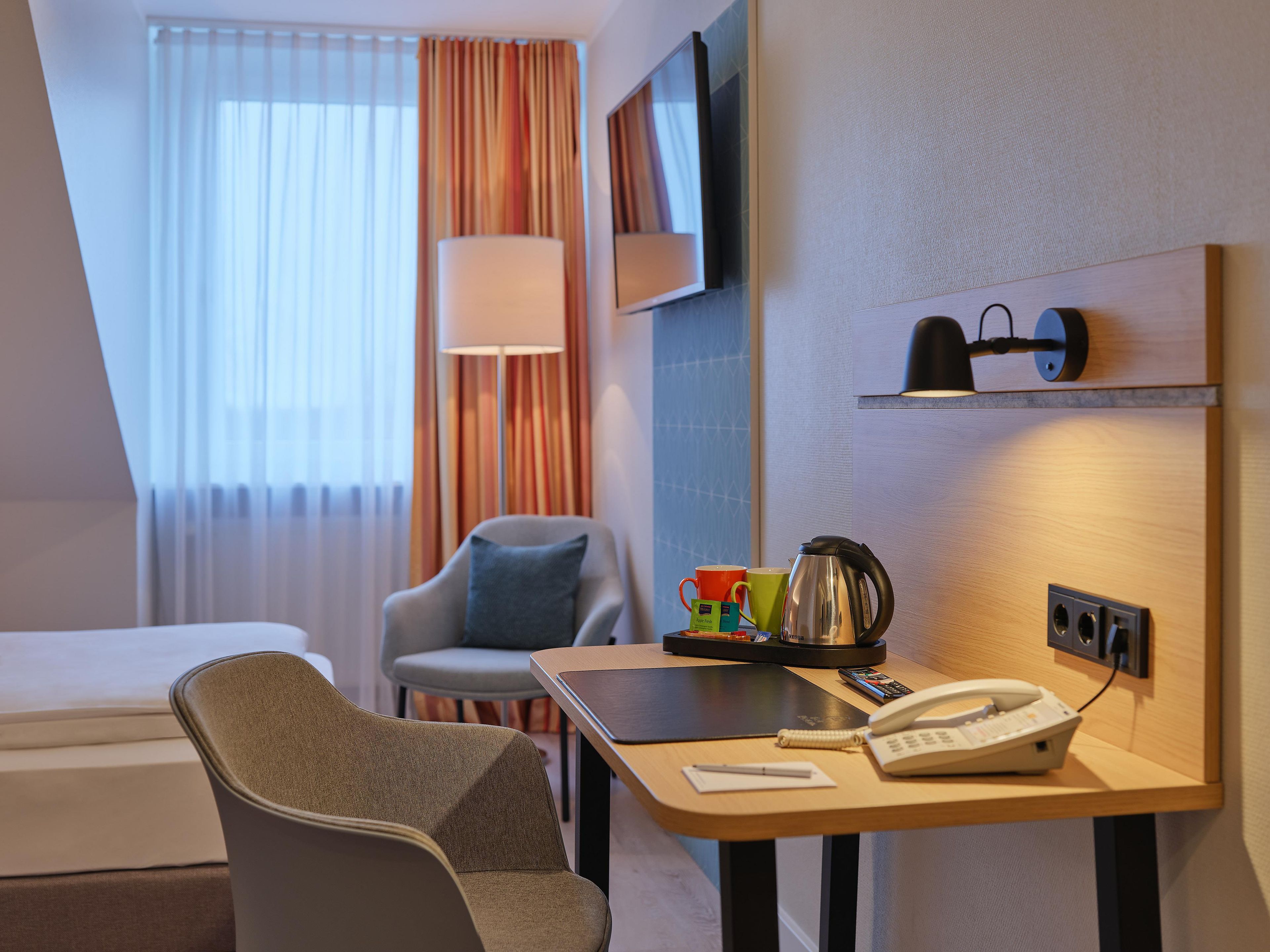 A hotel room with a bed, a desk with a telephone and a kettle, a chair, a TV, and a window with curtains.