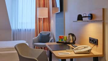 Habitación de hotel con cama, escritorio con teléfono y tetera, una silla, televisor y ventana con cortinas.
