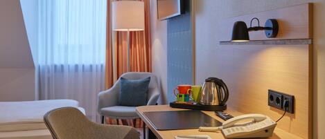 Habitación de hotel con cama, escritorio con teléfono y tetera, una silla, televisor y ventana con cortinas.