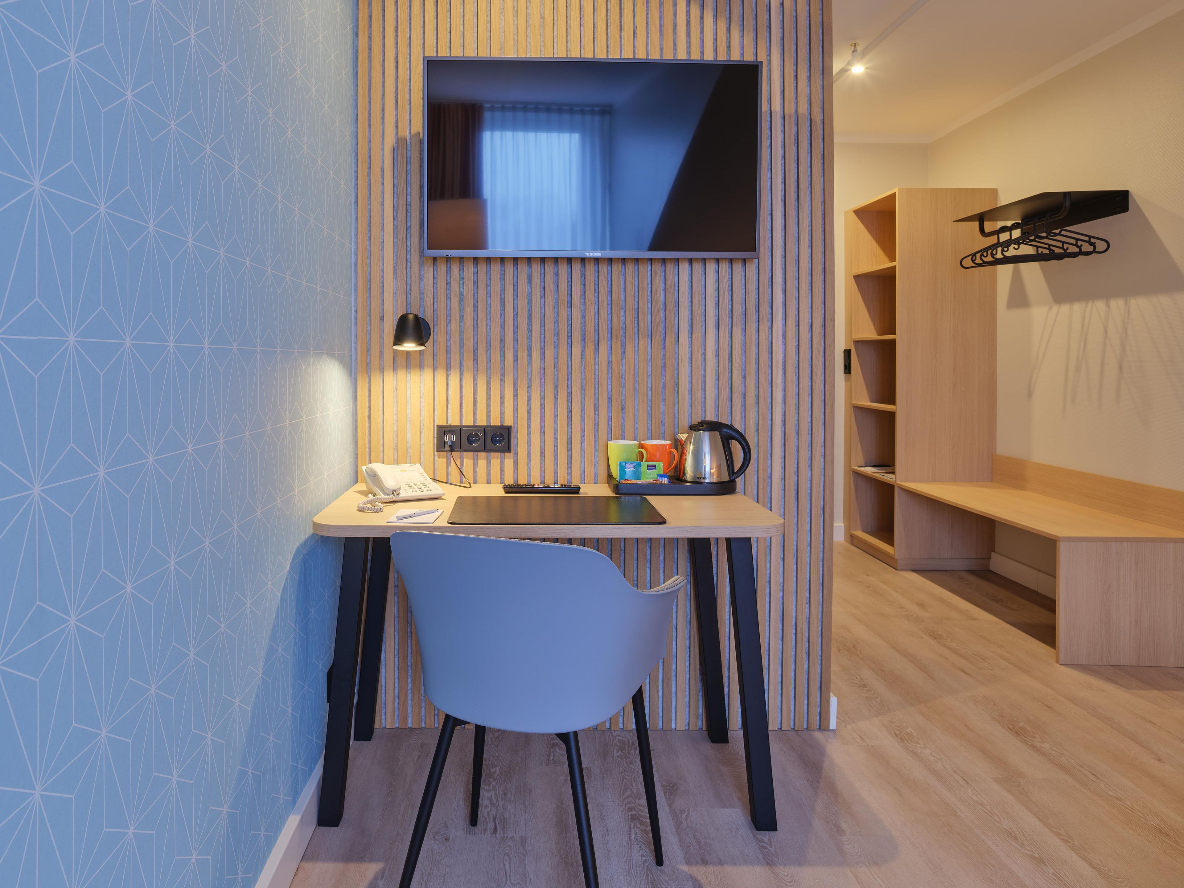 Une chambre moderne équipée d’un bureau, d’une chaise, d’une télévision et d’une étagère sur laquelle se trouve une bouilloire.