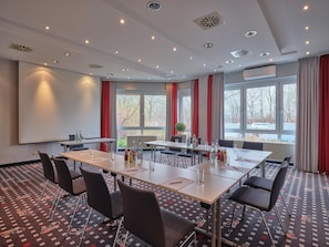 Een vergaderzaal met een grote tafel, stoelen, een projectiescherm en ramen voorzien van gordijnen.