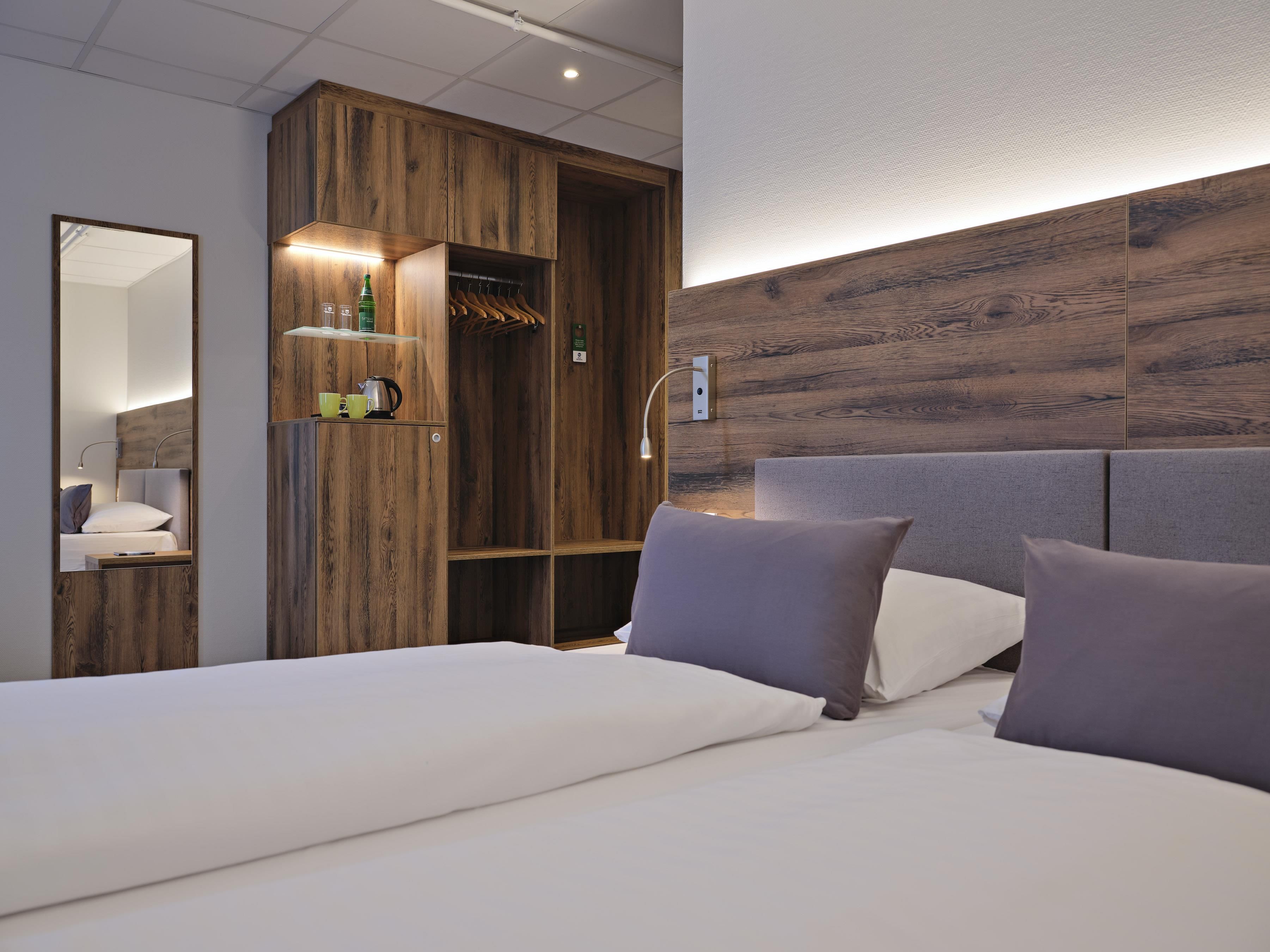 A modern hotel room with a large bed, a wooden wardrobe, and a mirror.