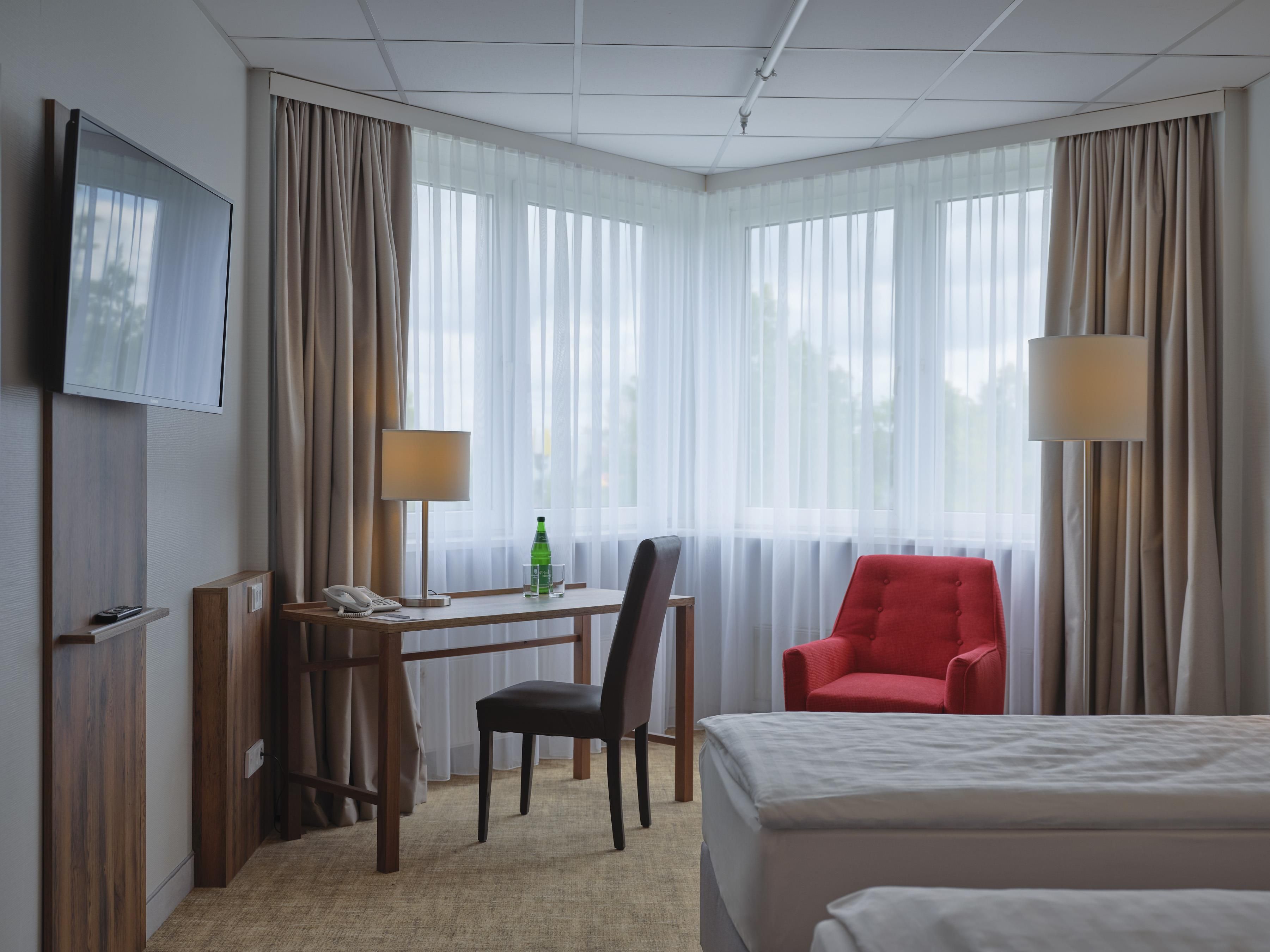Quarto de hotel com cama, escrivaninha com telefone, cadeira, televisão e luminária.