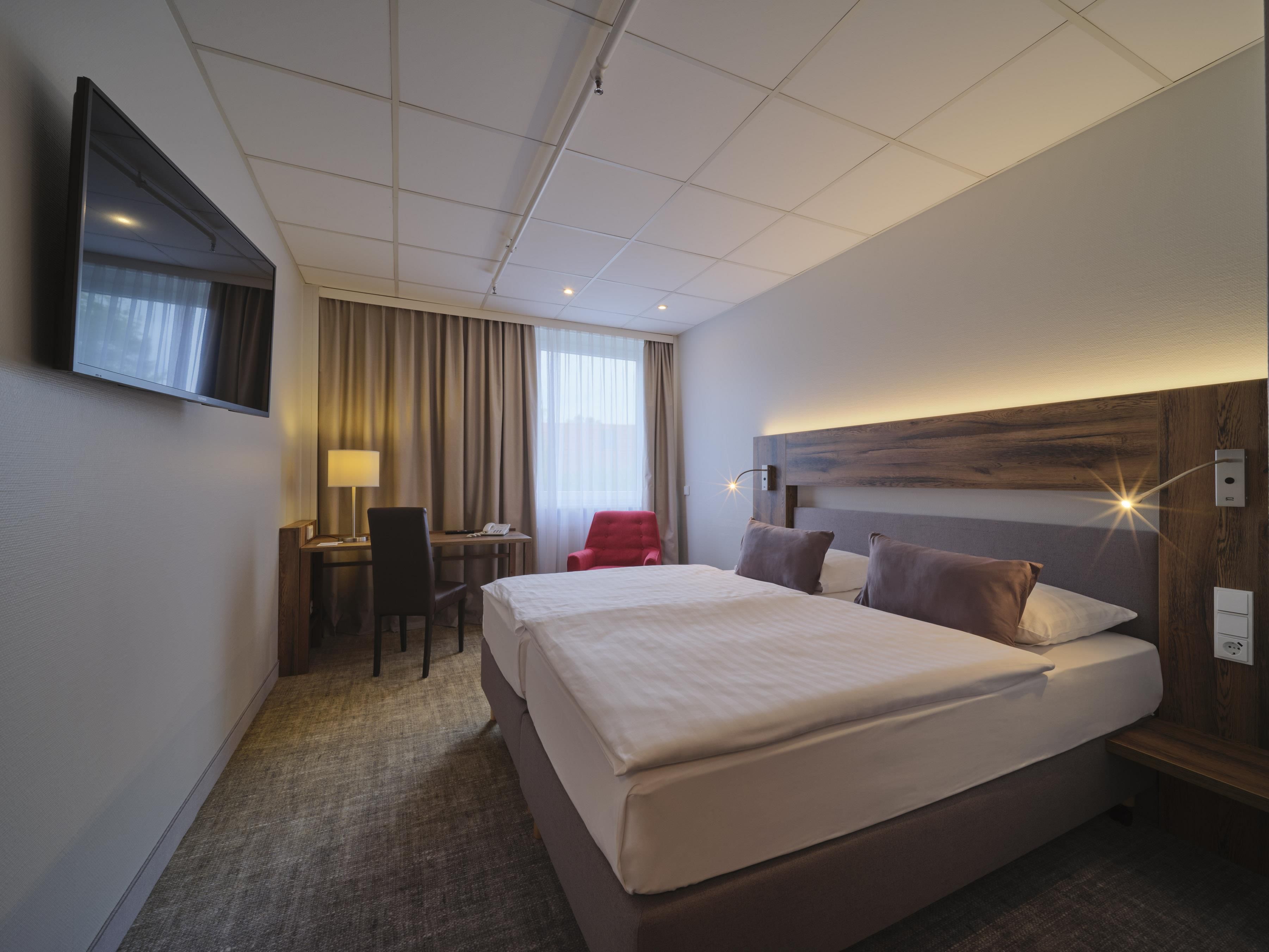 Hotellihuone, jossa on suuri sänky, työpöytä tuolilla, seinälle asennettu televisio ja ikkuna, jossa on verhot.
