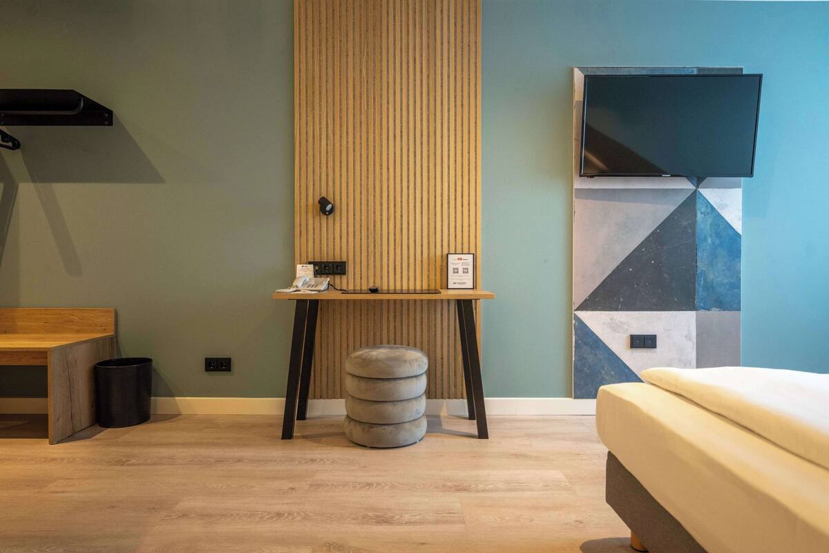 A modern hotel room with a wooden desk, a round ottoman, a bed, and a TV mounted on the wall.