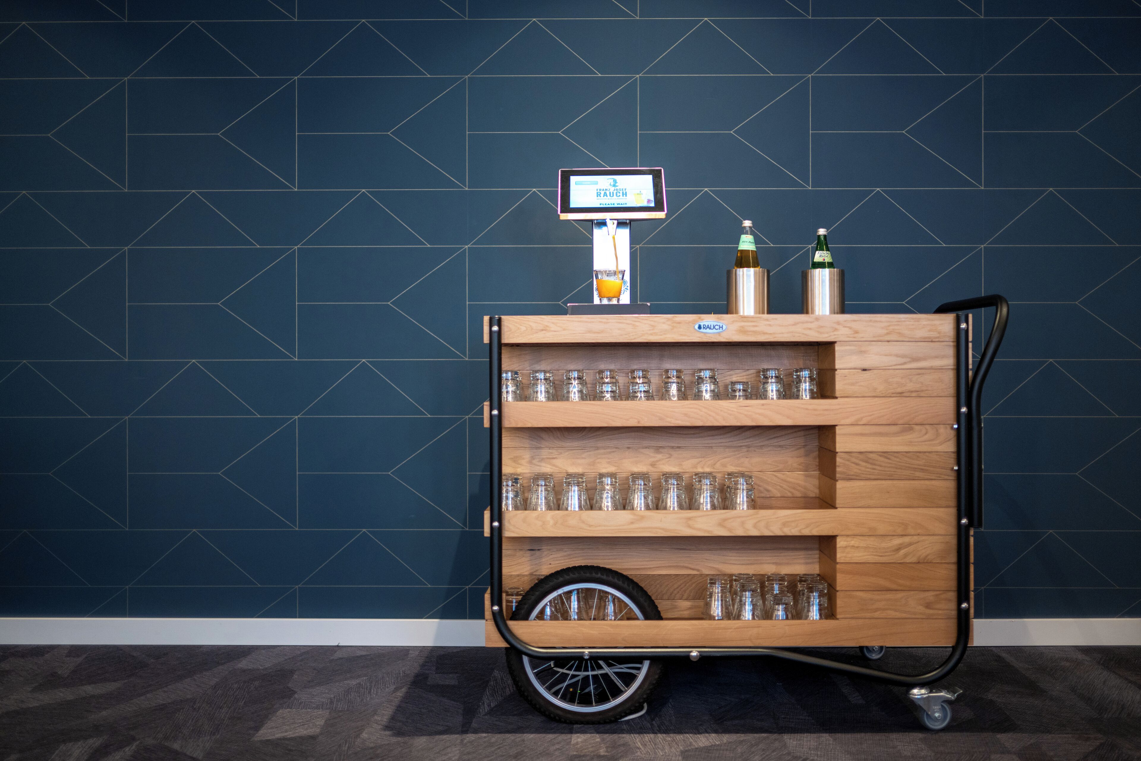 Un carro de madera con estantes que contiene vasos y botellas, frente a una pared de azulejos azul oscuro.