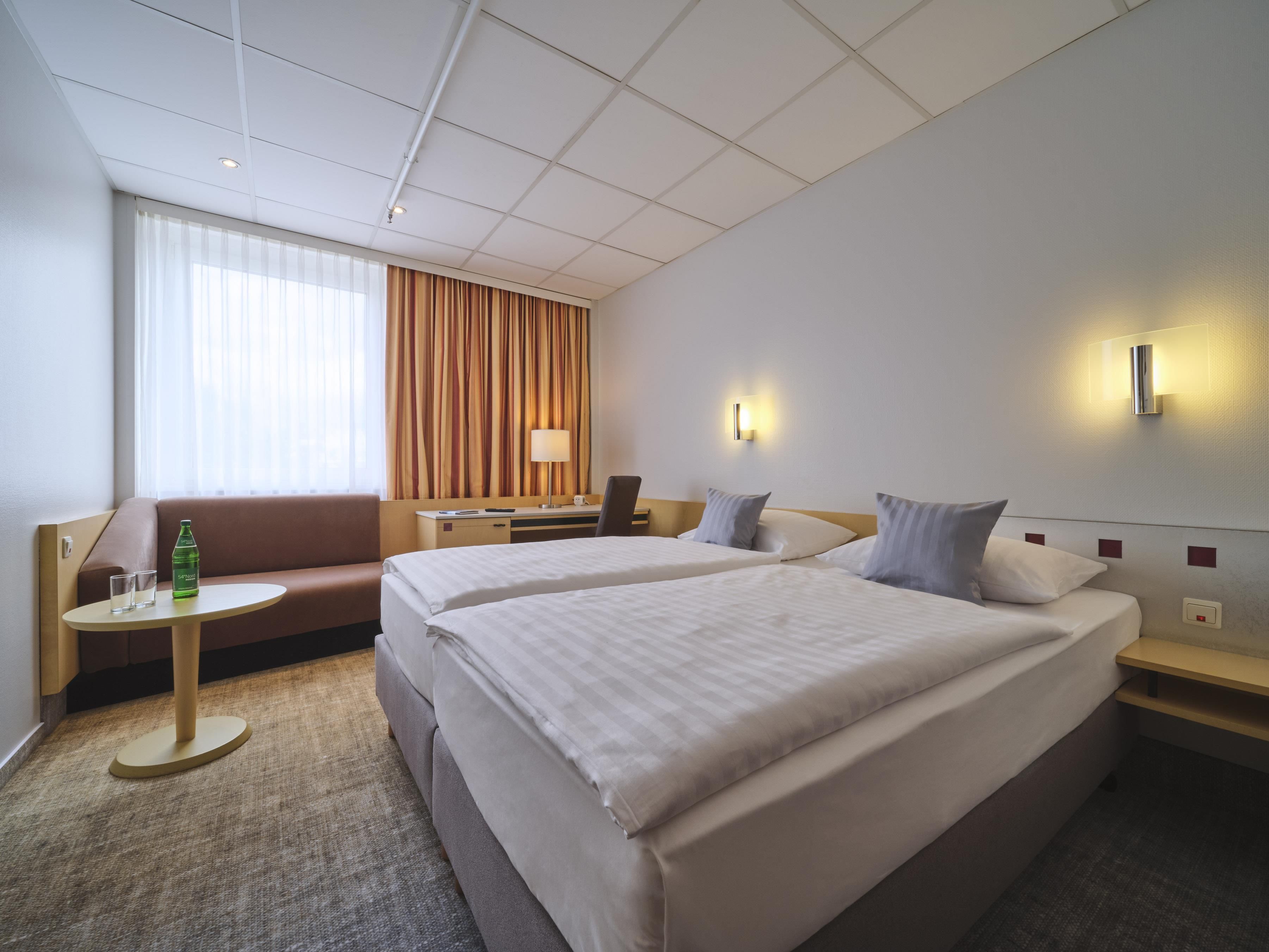 A hotel room with two beds, a sofa, a small table, a lamp, and a window with curtains.