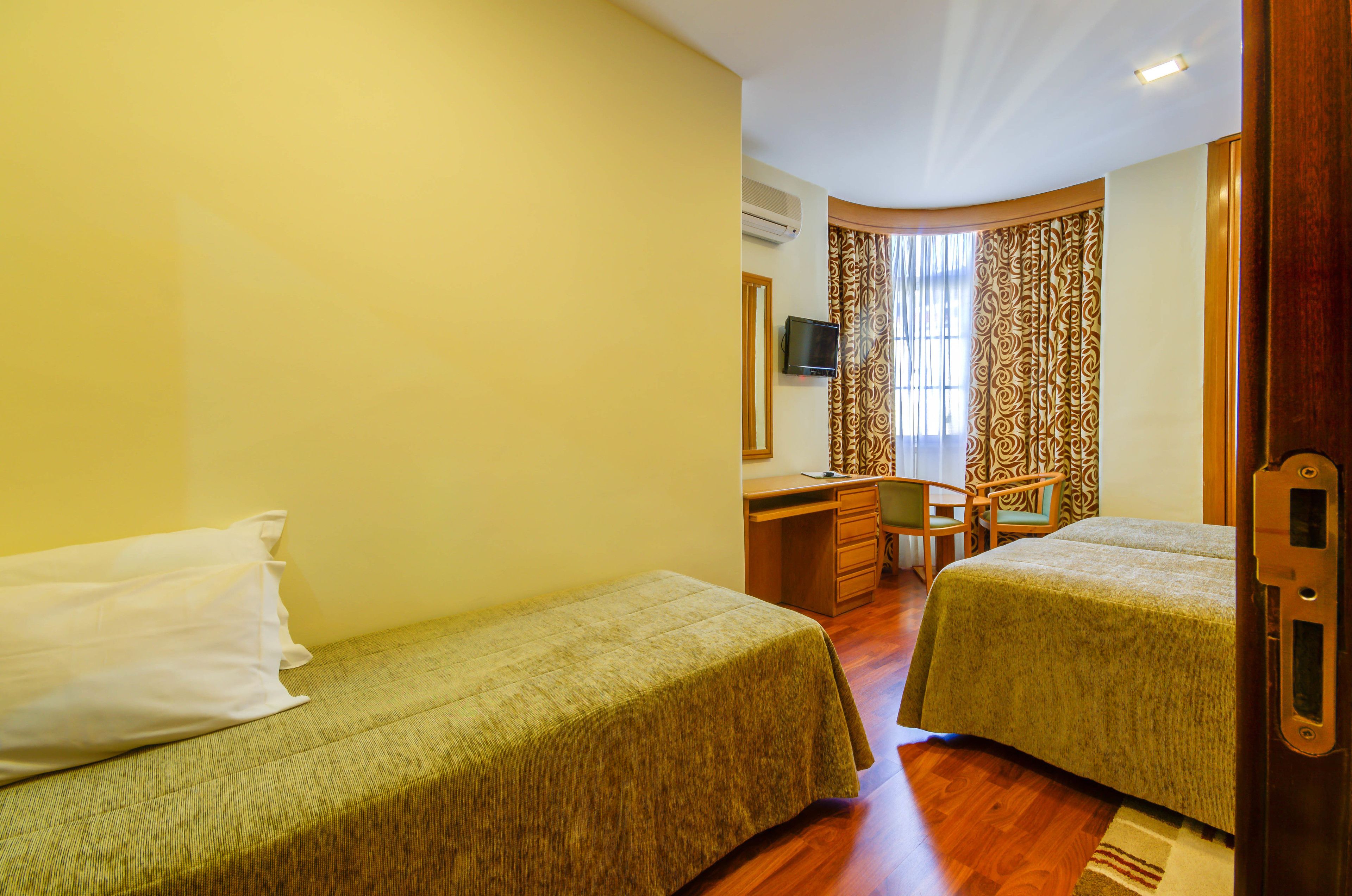 A hotel room with two single beds, a desk with a chair, a television, and a window with curtains.