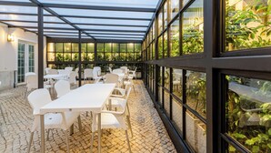 A covered outdoor area with tables and chairs, large windows, and a view of greenery. - Hotel Avenida Park (Lisbon)