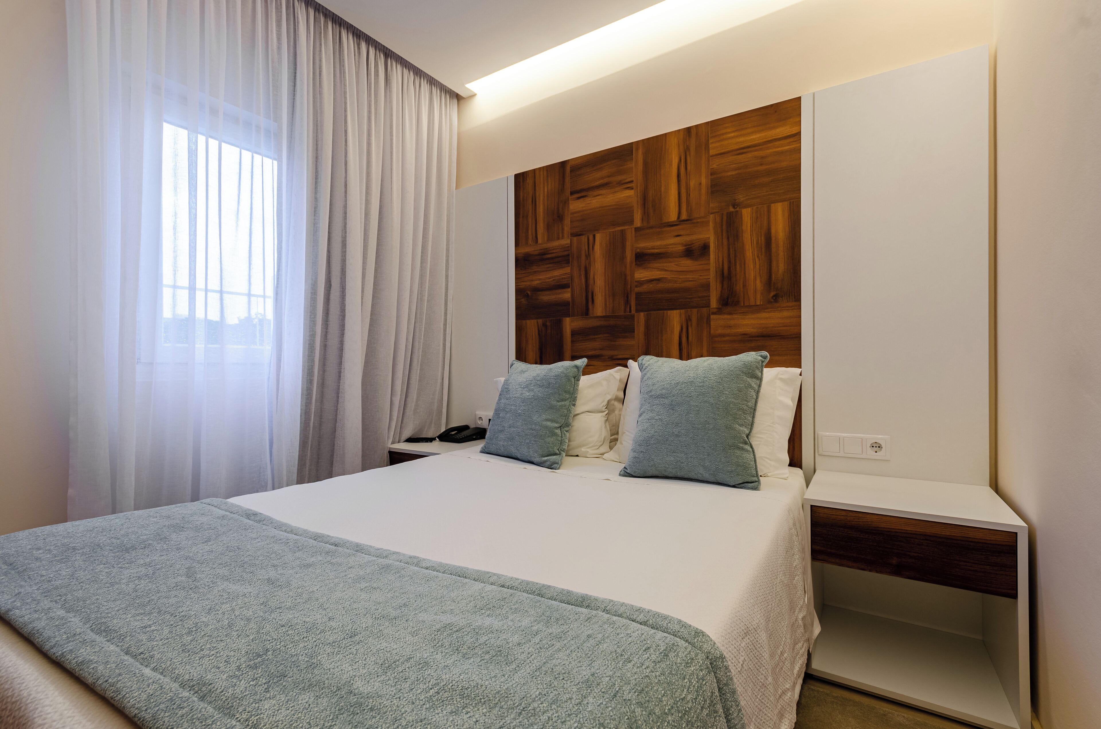 A hotel room with a bed, a wooden headboard, a bedside table, and a window with curtains.