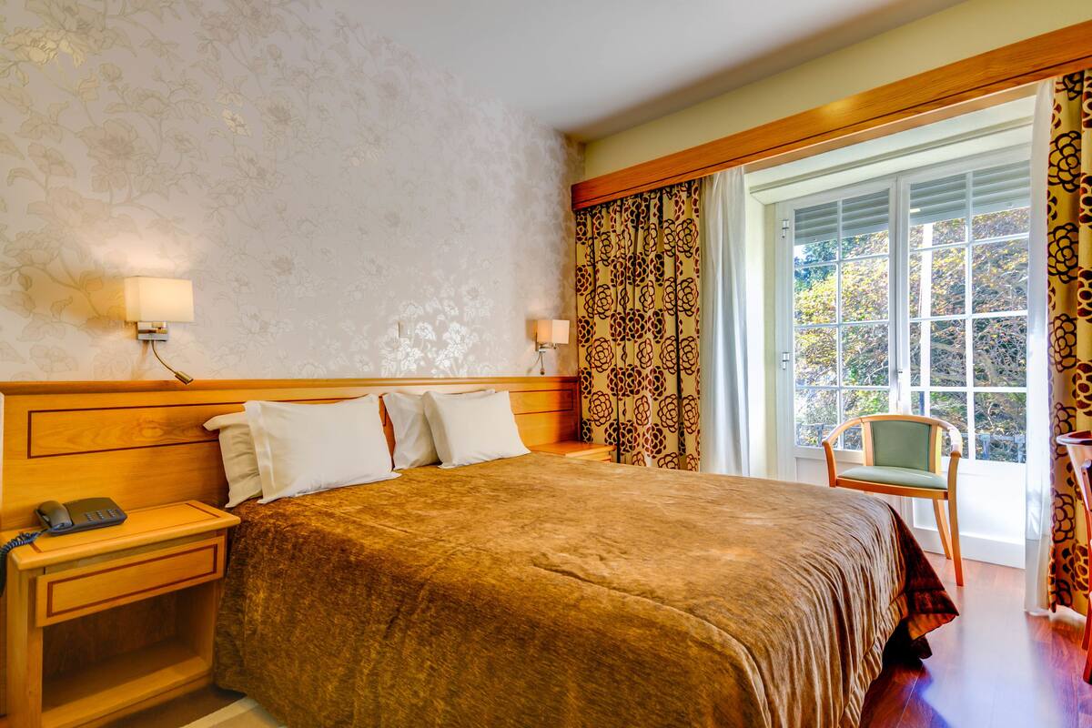 Een hotelkamer met een groot bed, een houten hoofdbord, een nachtkastje met een lamp en een raam met gordijnen.