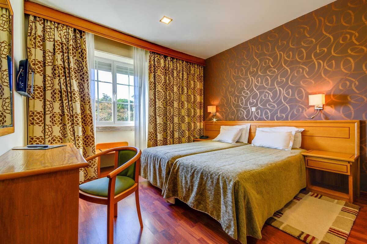 Een hotelkamer met twee bedden, een bureau, een stoel, een raam met gordijnen, een behang met patroon en een nachtkastje met een lamp.