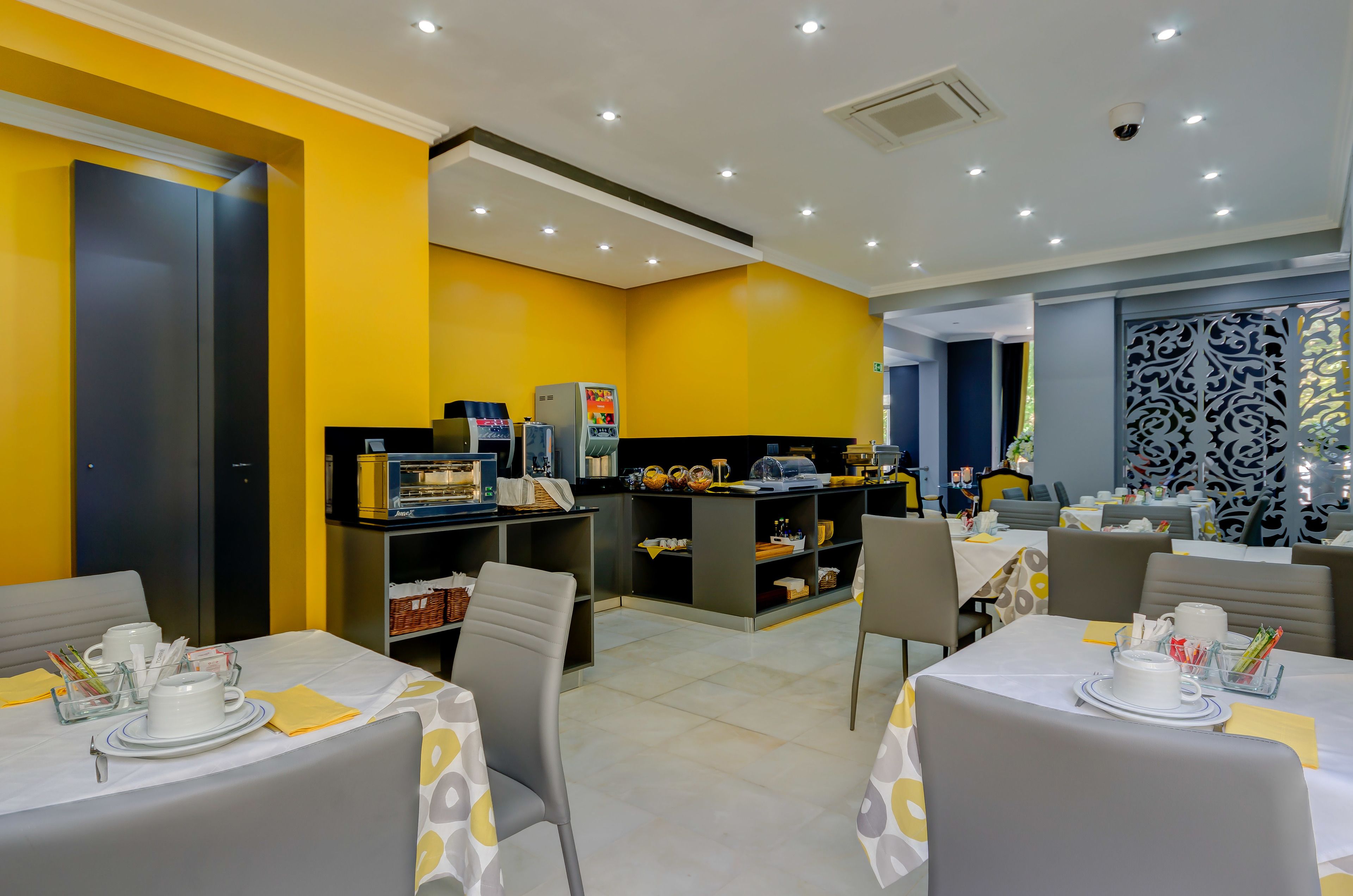 A brightly lit dining area with yellow walls, a kitchen area with appliances, and tables set with white tablecloths and napkins.