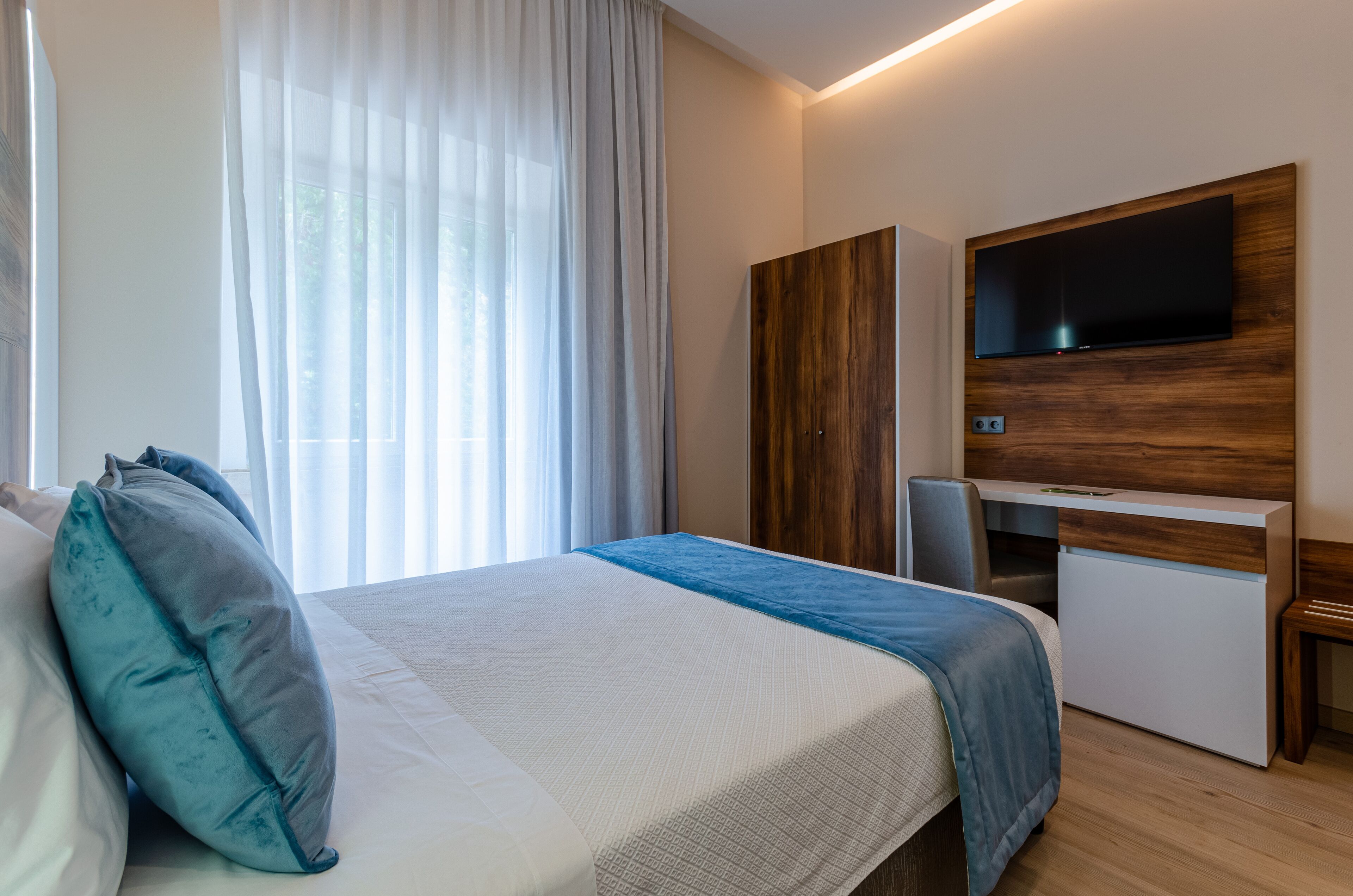 Habitación de hotel con cama, televisor montado en un panel de madera y ventana con cortinas traslúcidas.