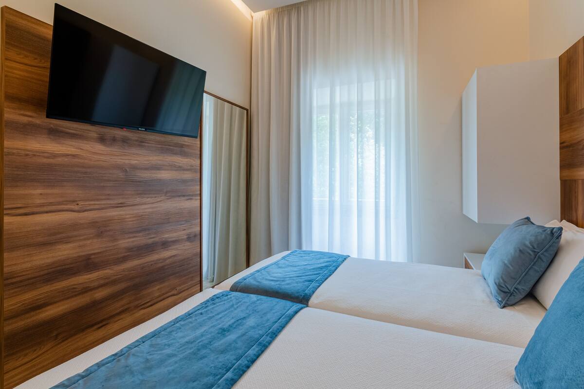 Een moderne hotelkamer met een groot bed, een flatscreen televisie bevestigd aan een houten paneel en een groot raam met dunne gordijnen.