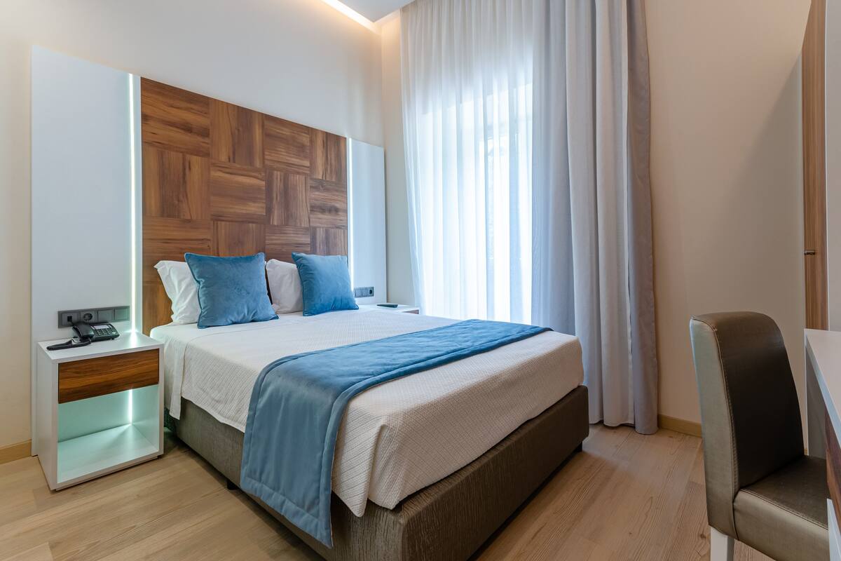 Een moderne hotelkamer met een houten hoofdeinde, een groot bed met blauw beddengoed, een nachtkastje met telefoon en een stoel.