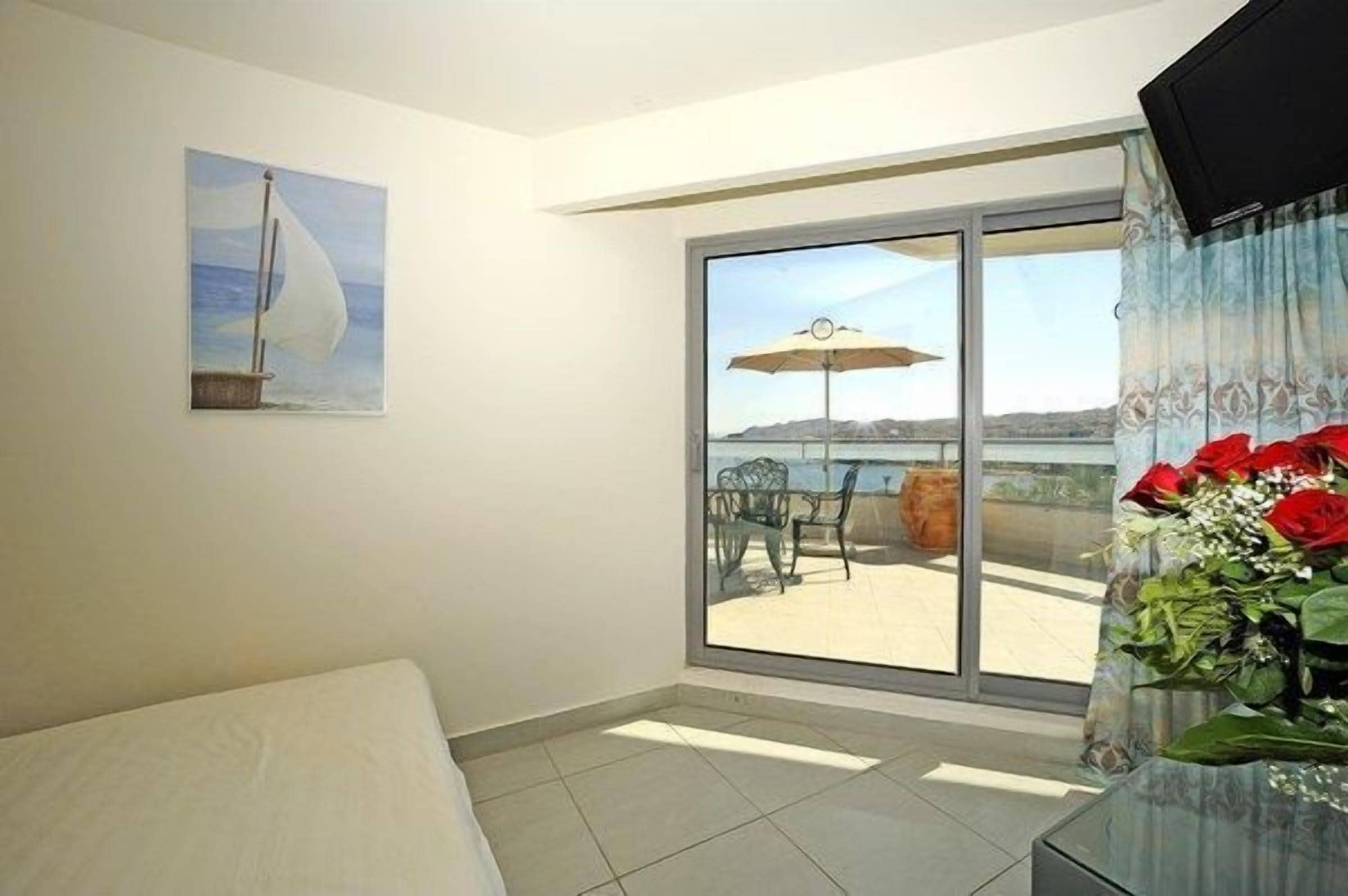 A room with a bed, a painting of a sailboat, a balcony with a table and chairs, and a large window.