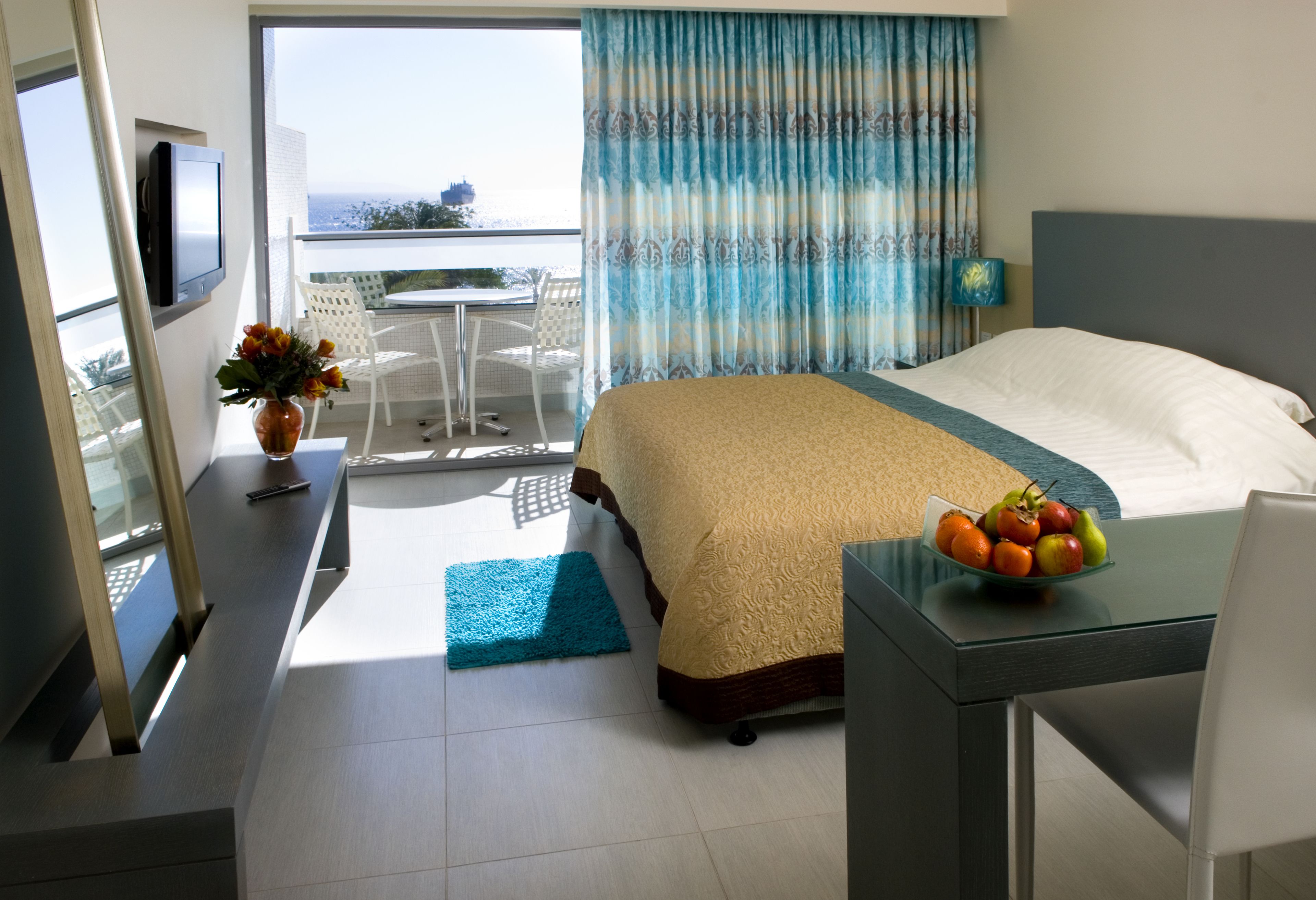 Habitación de hotel con cama, escritorio con frutero, televisión y balcón con vistas.