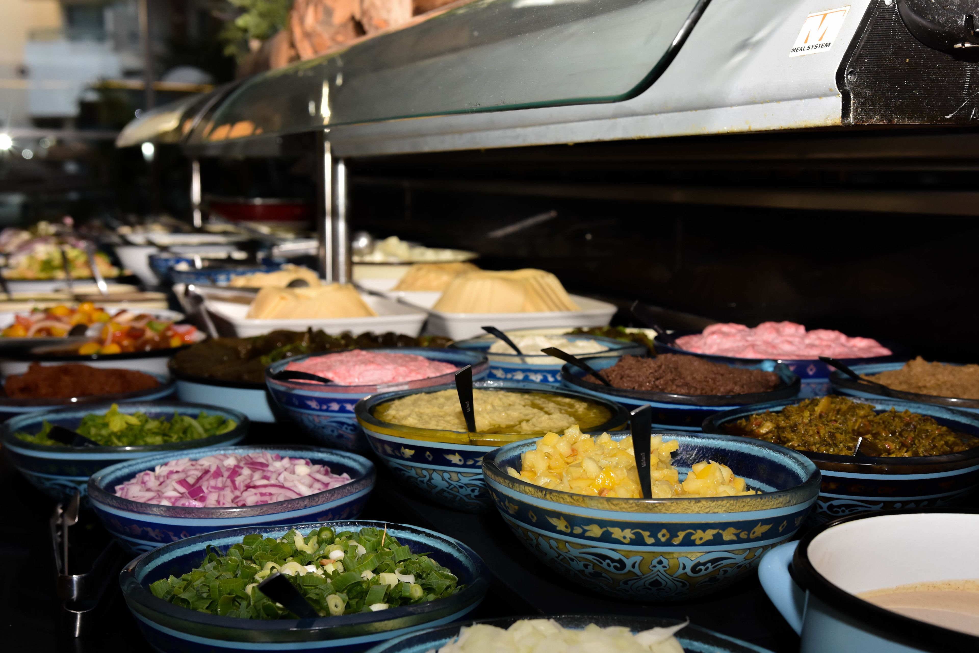Un bufé con diversos platos en recipientes azules y blancos, entre ellos ensaladas, carnes y otros alimentos ya preparados.