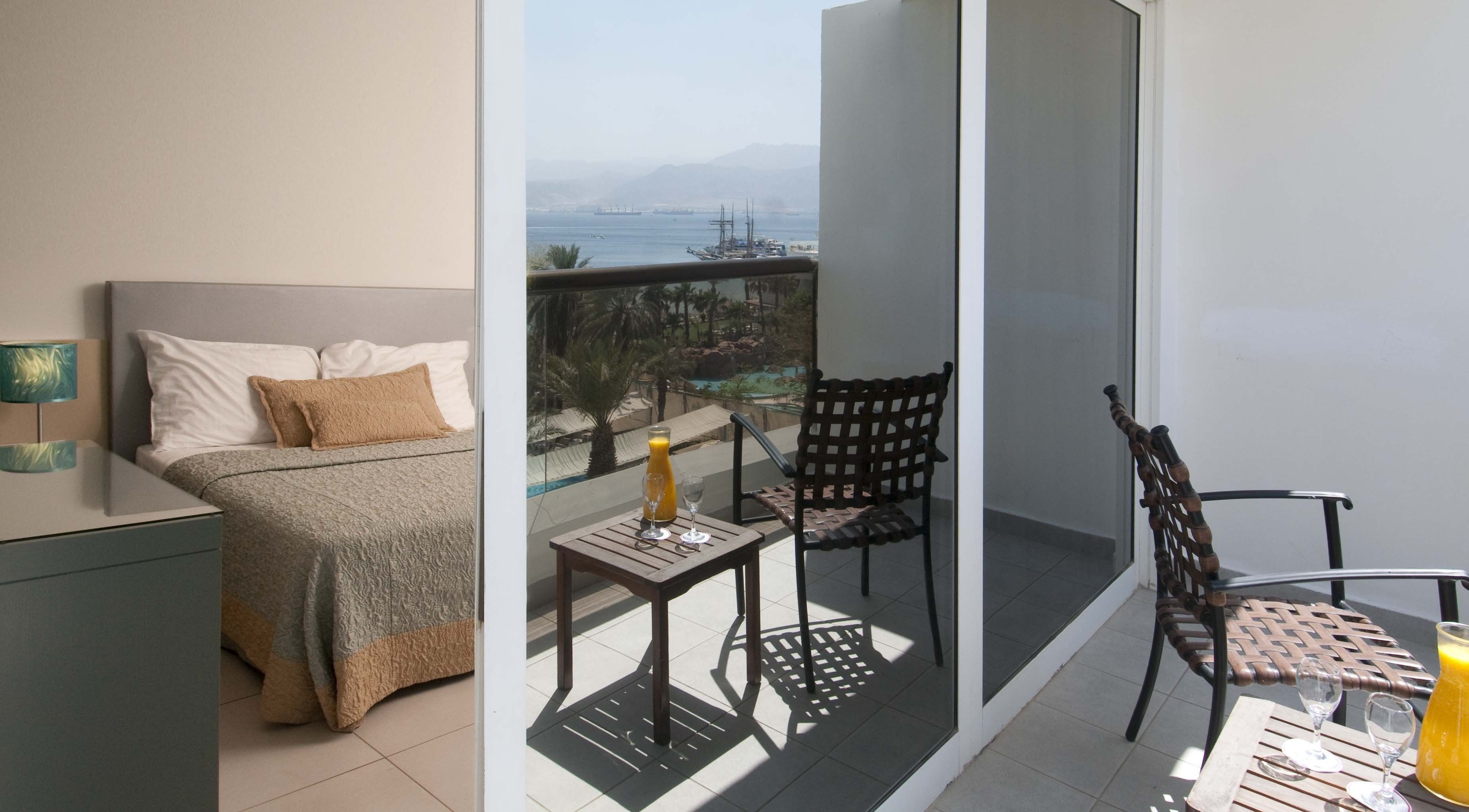 Habitación de hotel con una cama, una mesita con una bebida y un balcón con vistas al mar y a las embarcaciones.