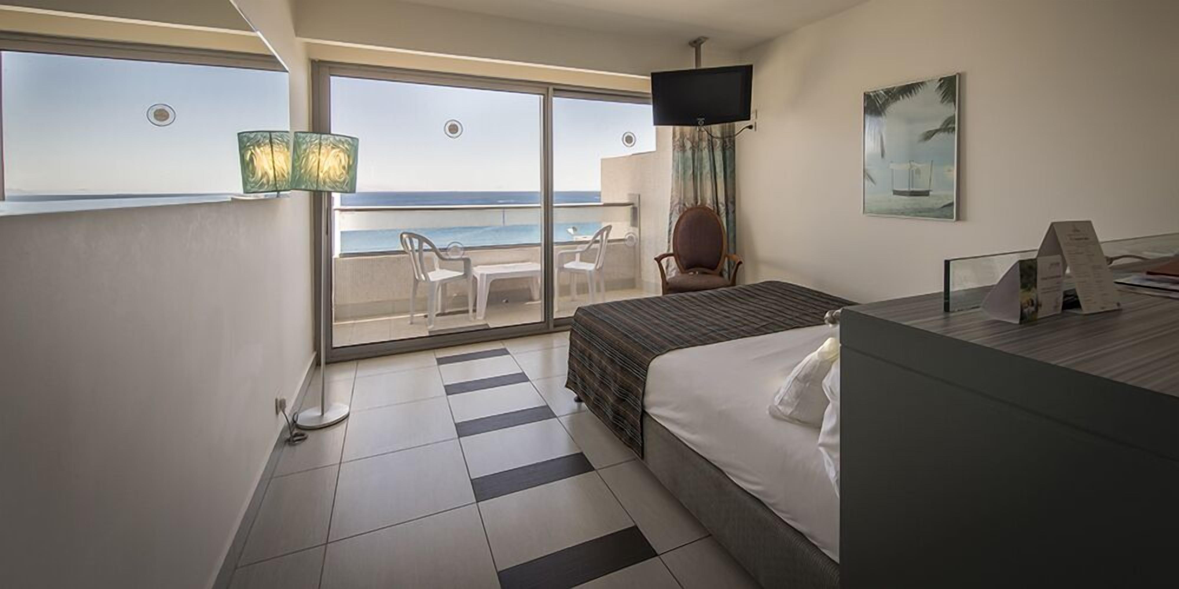 Habitación de hotel con cama, televisión, escritorio y balcón con vista al mar.