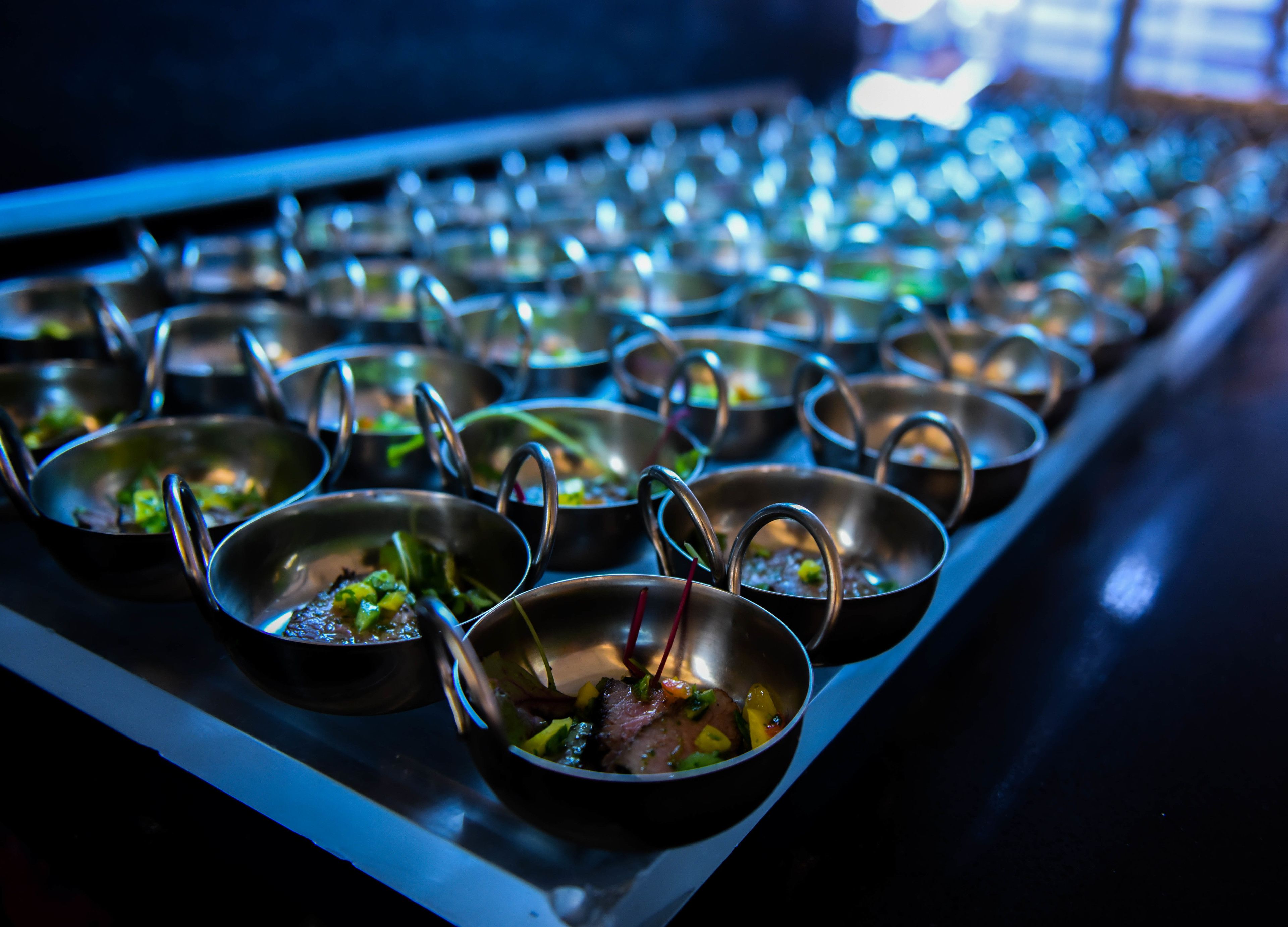 A row of metal bowls with handles, each containing food, arranged on a tray.