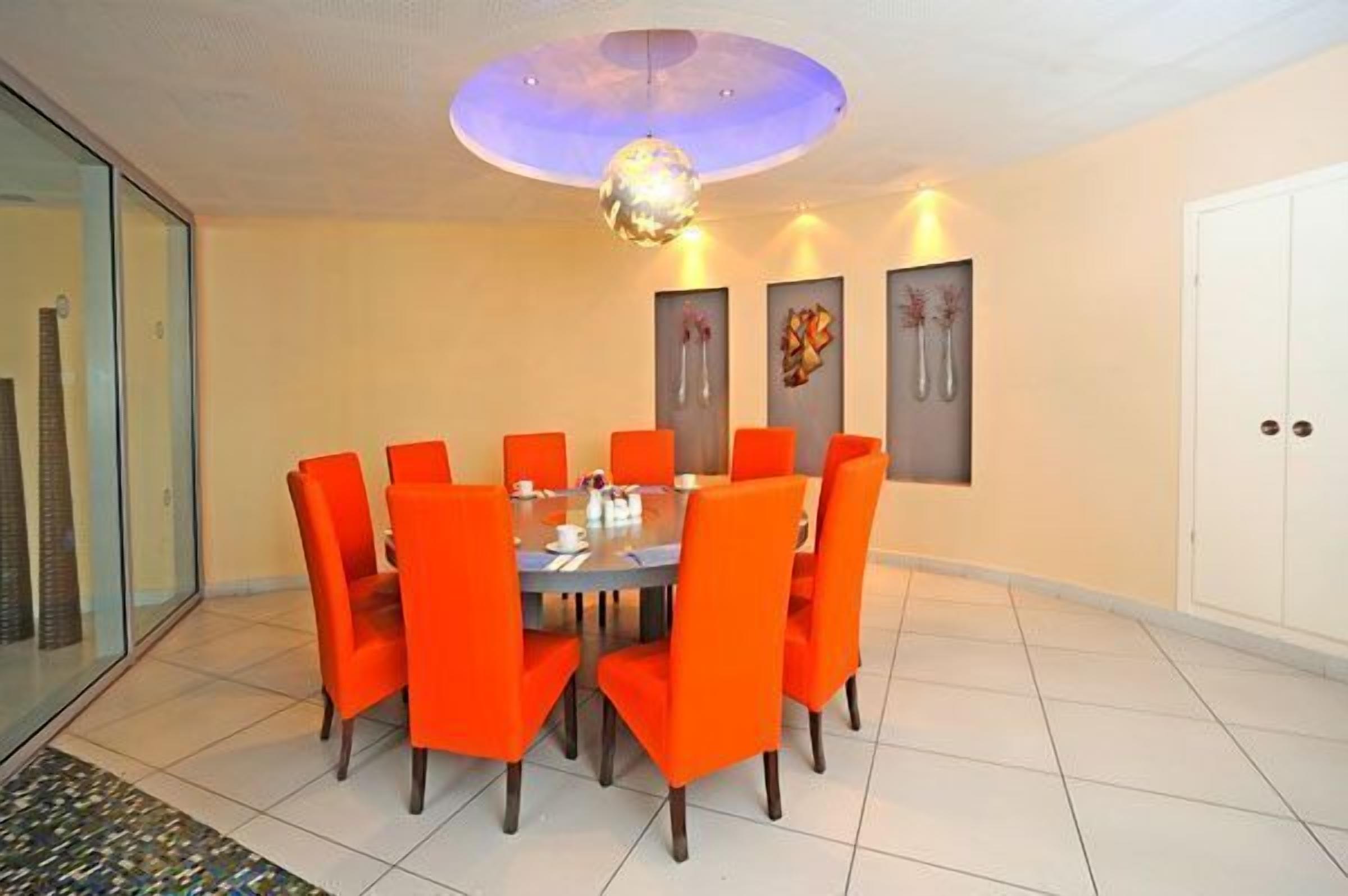 A dining area with a round table and orange chairs, a glass partition, and a ceiling with a spherical light fixture.