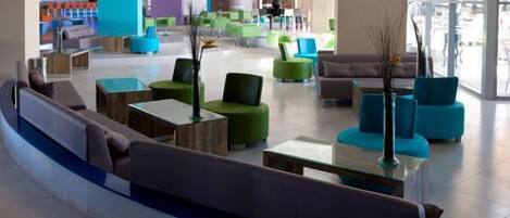 A modern lobby with green and blue seating, a central coffee table, and a view of the outside through large windows.
