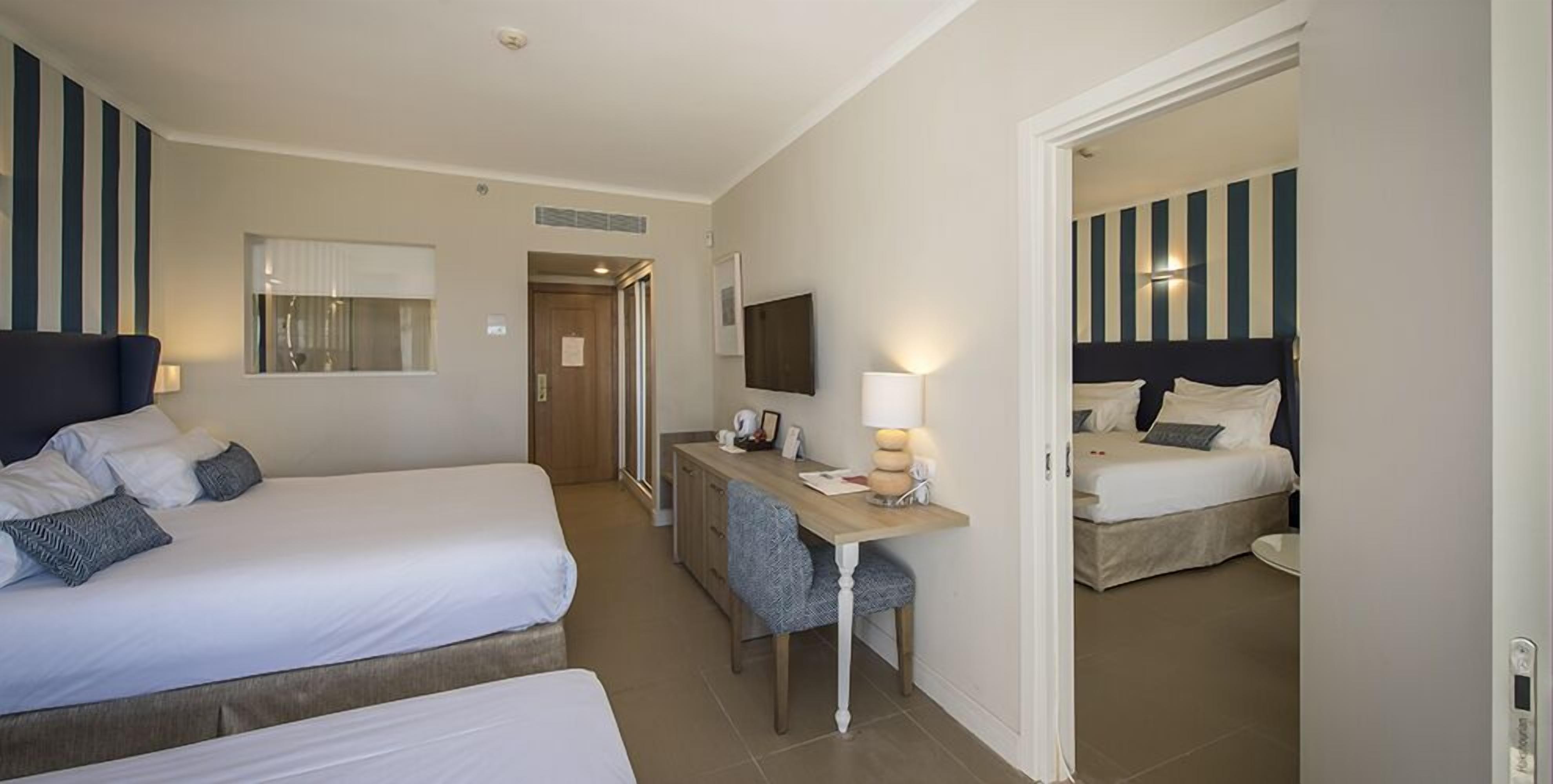 A hotel room with two beds, a desk with a lamp, a TV, and a door leading to another room.