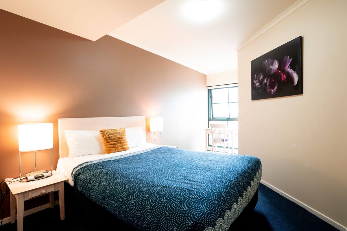 Quarto de hotel com uma cama grande, duas luminárias de cabeceira, mesa de apoio e uma janela.