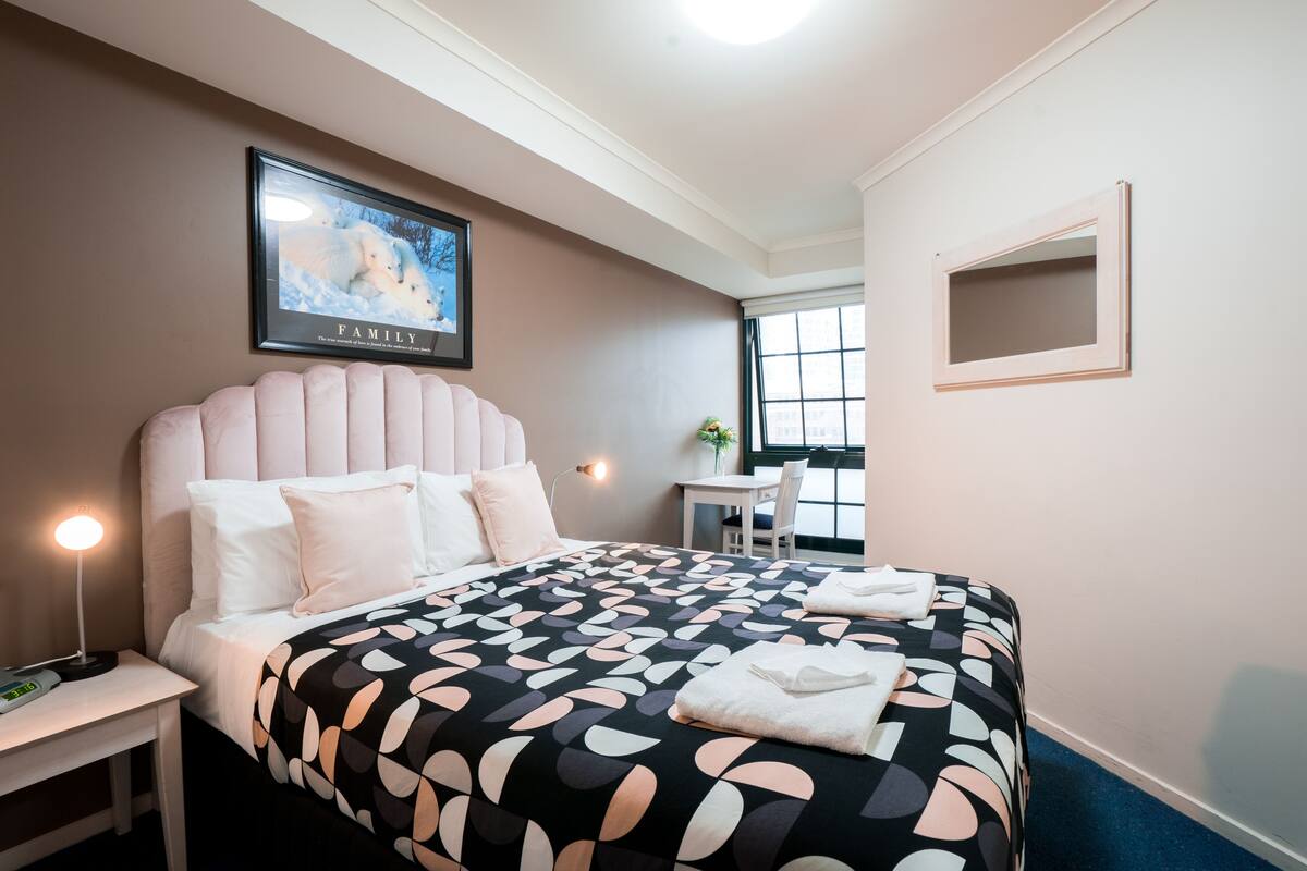 Quarto de hotel com uma cama grande, uma mesinha de cabeceira com abajur, um quadro na parede e uma janela com vista.
