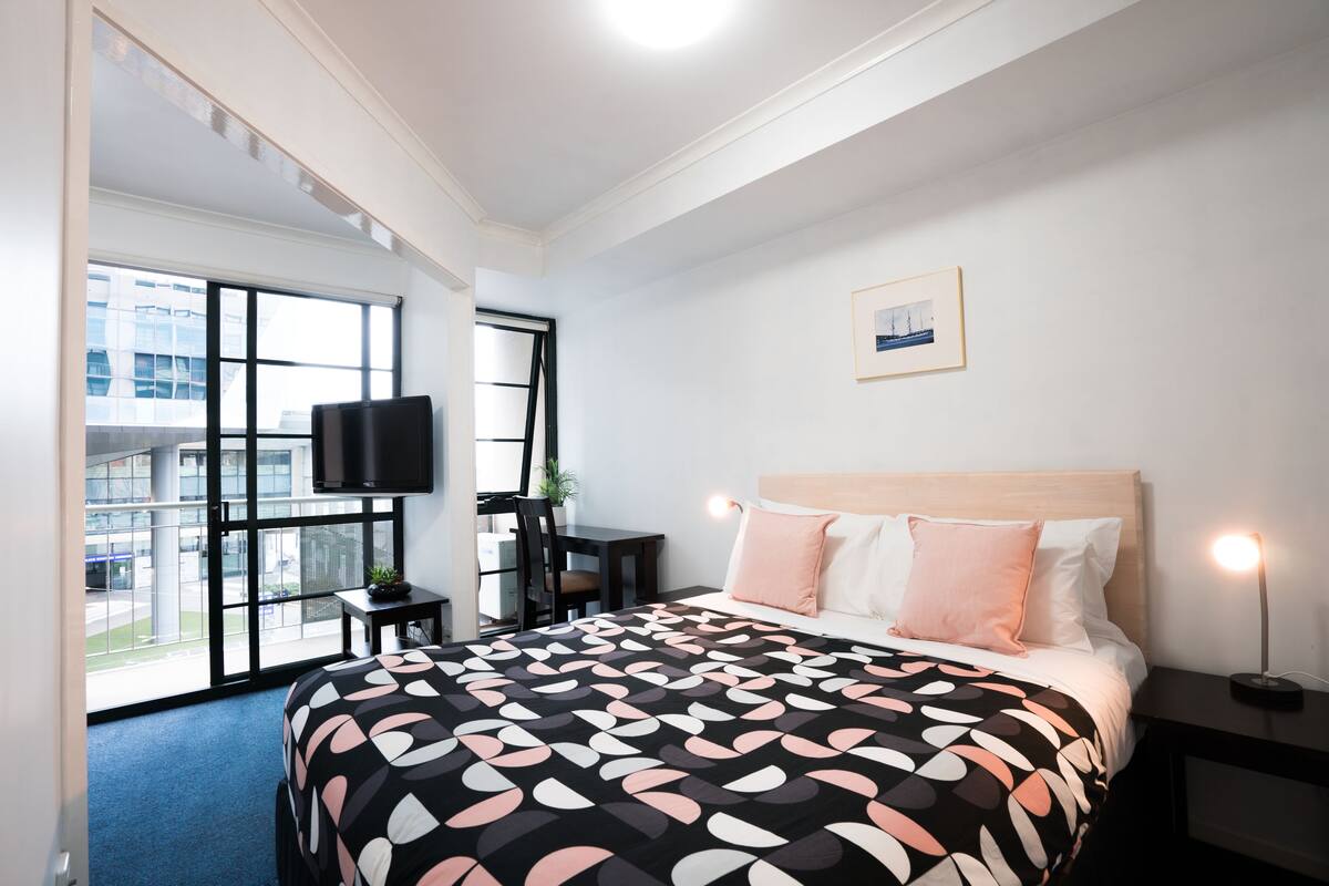 Quarto de hotel moderno com uma cama grande, televisão, escrivaninha e janela com vista para a cidade.