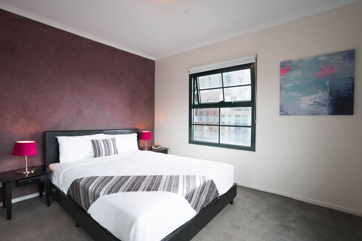 Quarto de hotel com uma cama grande, mesa de cabeceira com abajur, janela com vista para prédios e um quadro na parede.