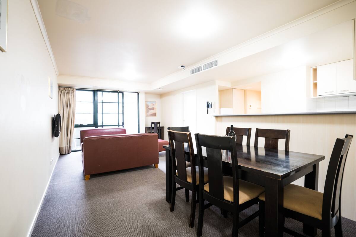 Uma sala de jantar com mesa, cadeiras, um sofá e uma janela com cortinas.