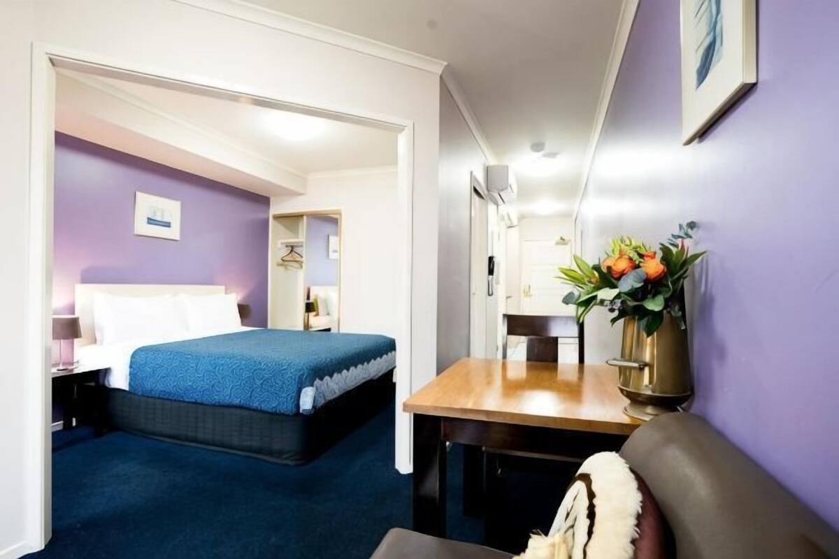 Quarto de hotel com cama, escrivaninha, cadeira, mesa pequena e um vaso com flores.