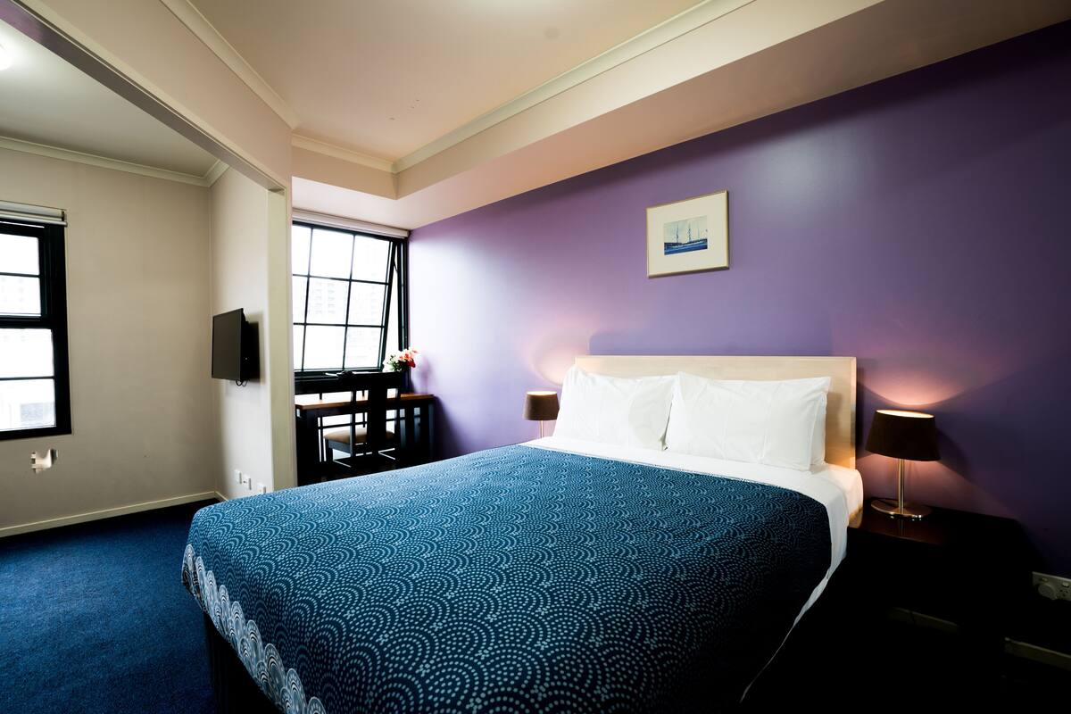 Quarto de hotel com uma cama grande, uma escrivaninha, uma cadeira, uma janela e uma televisão fixada na parede.