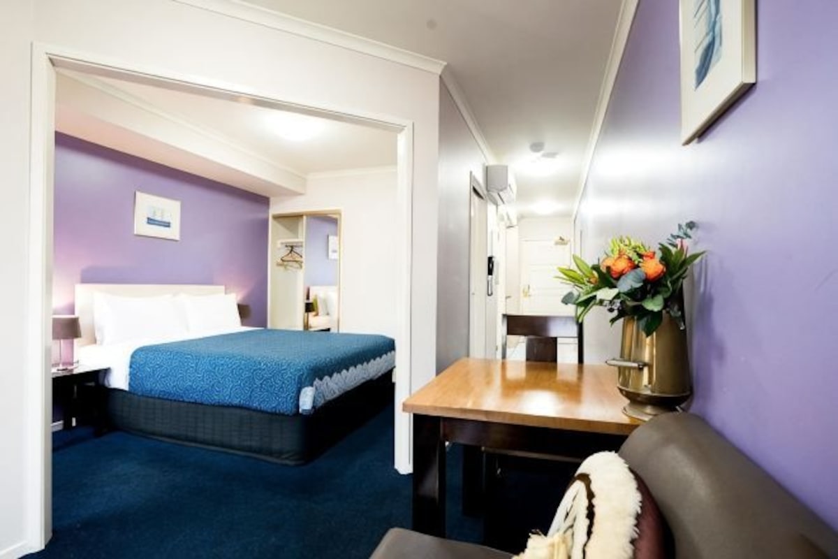 A hotel room with a bed, a desk, a chair, a small table, and a vase with flowers.