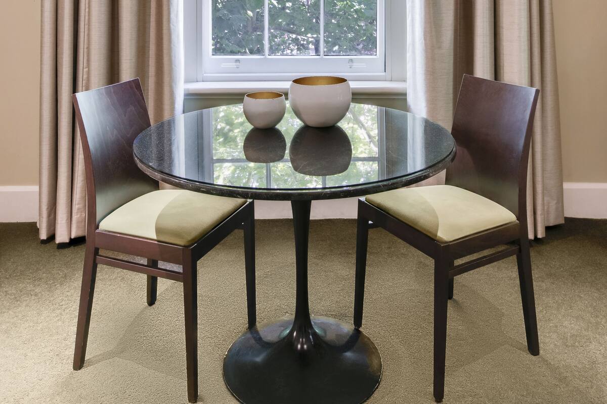 A room with a round glass table, two chairs, and a window with curtains.