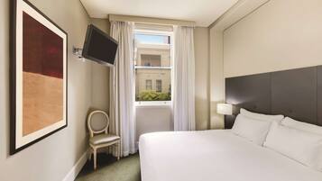 Habitación de hotel con cama, televisión montada en la pared, una silla, ventana con cortinas y vista al edificio exterior.
