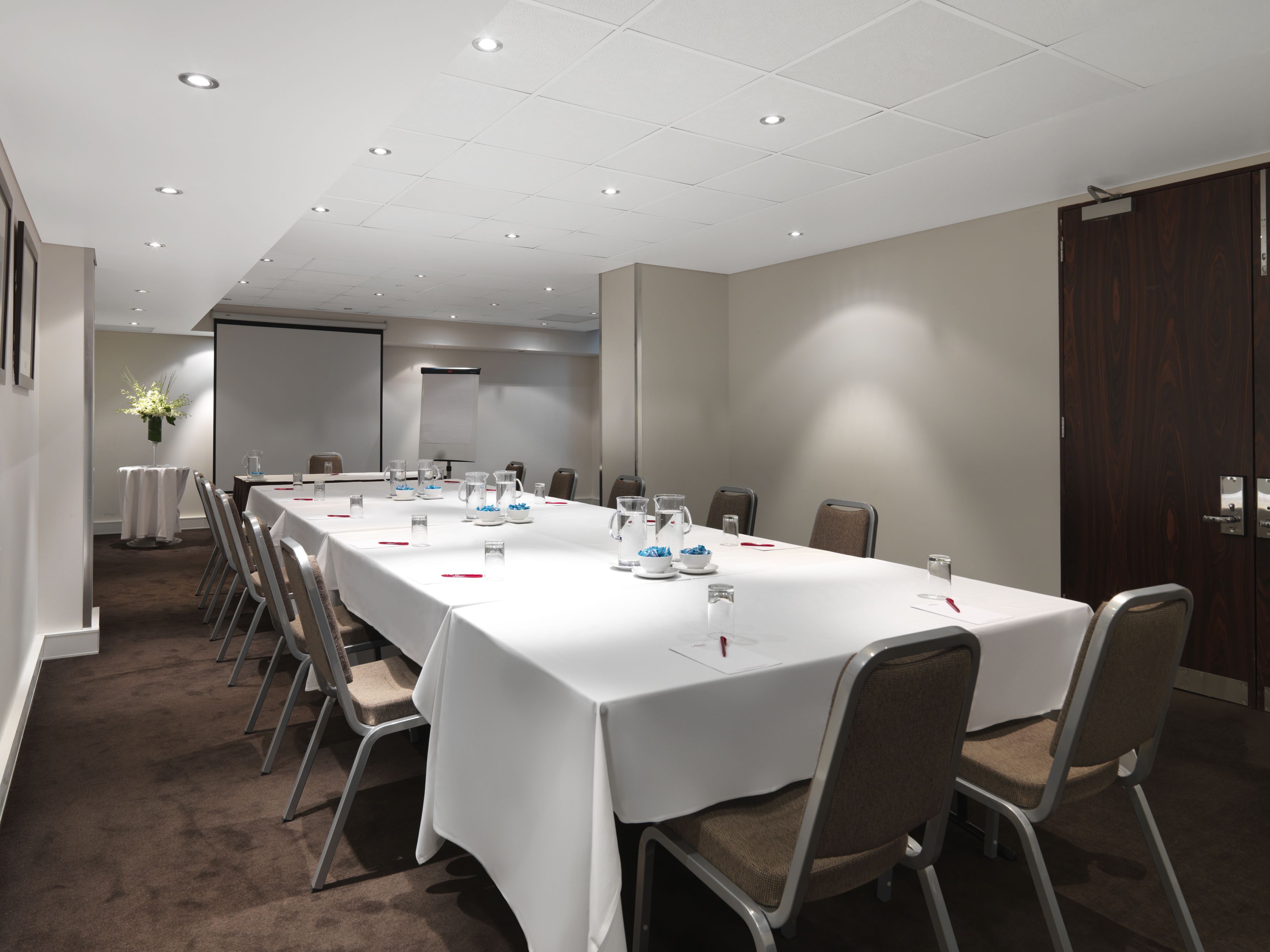 Un salón de conferencias con una larga mesa, sillas, una pantalla de proyector y una pizarra.
