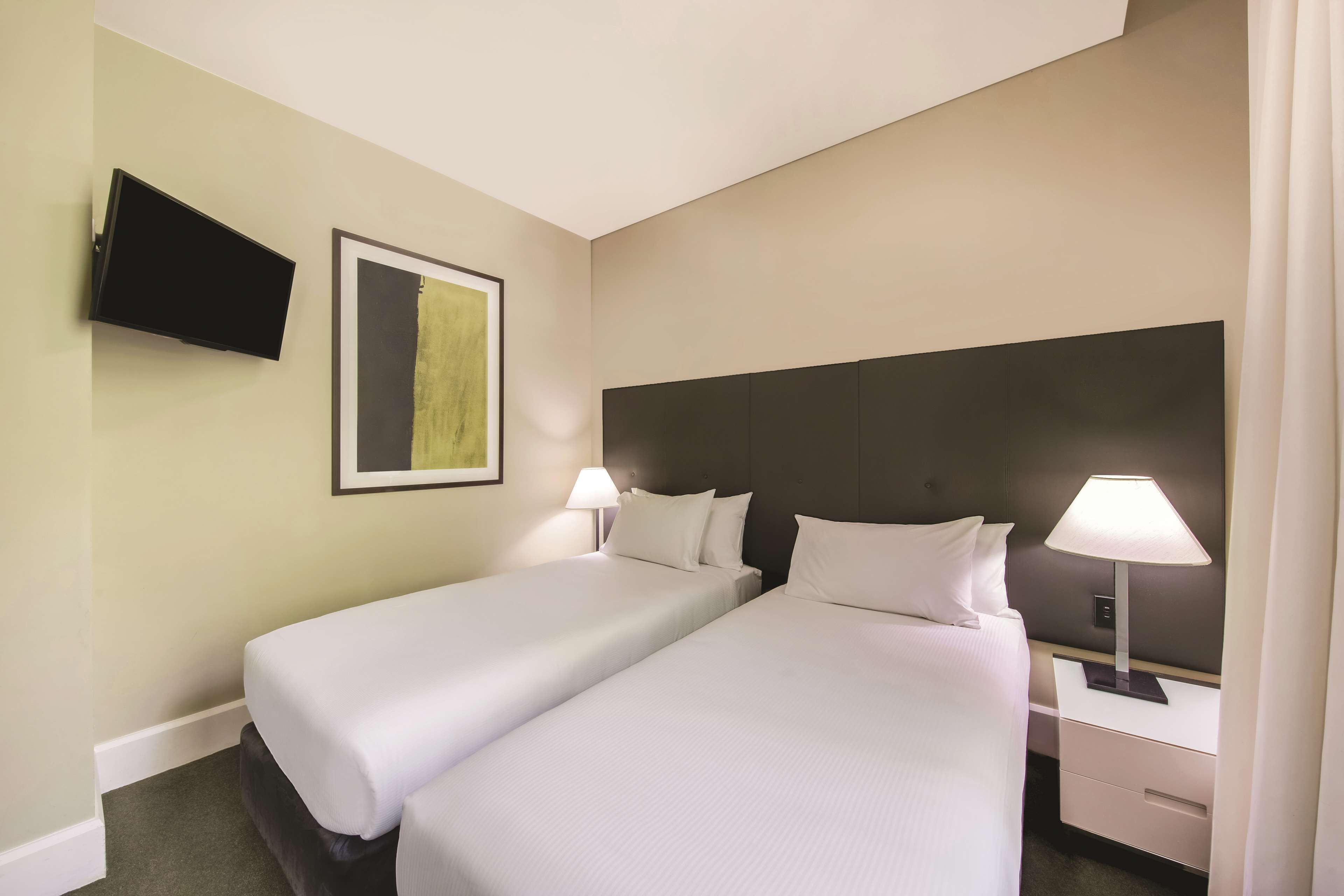 Habitación de hotel con dos camas, un televisor de pantalla plana montado en la pared, una mesita de noche con lámpara y una obra de arte enmarcada en la pared.