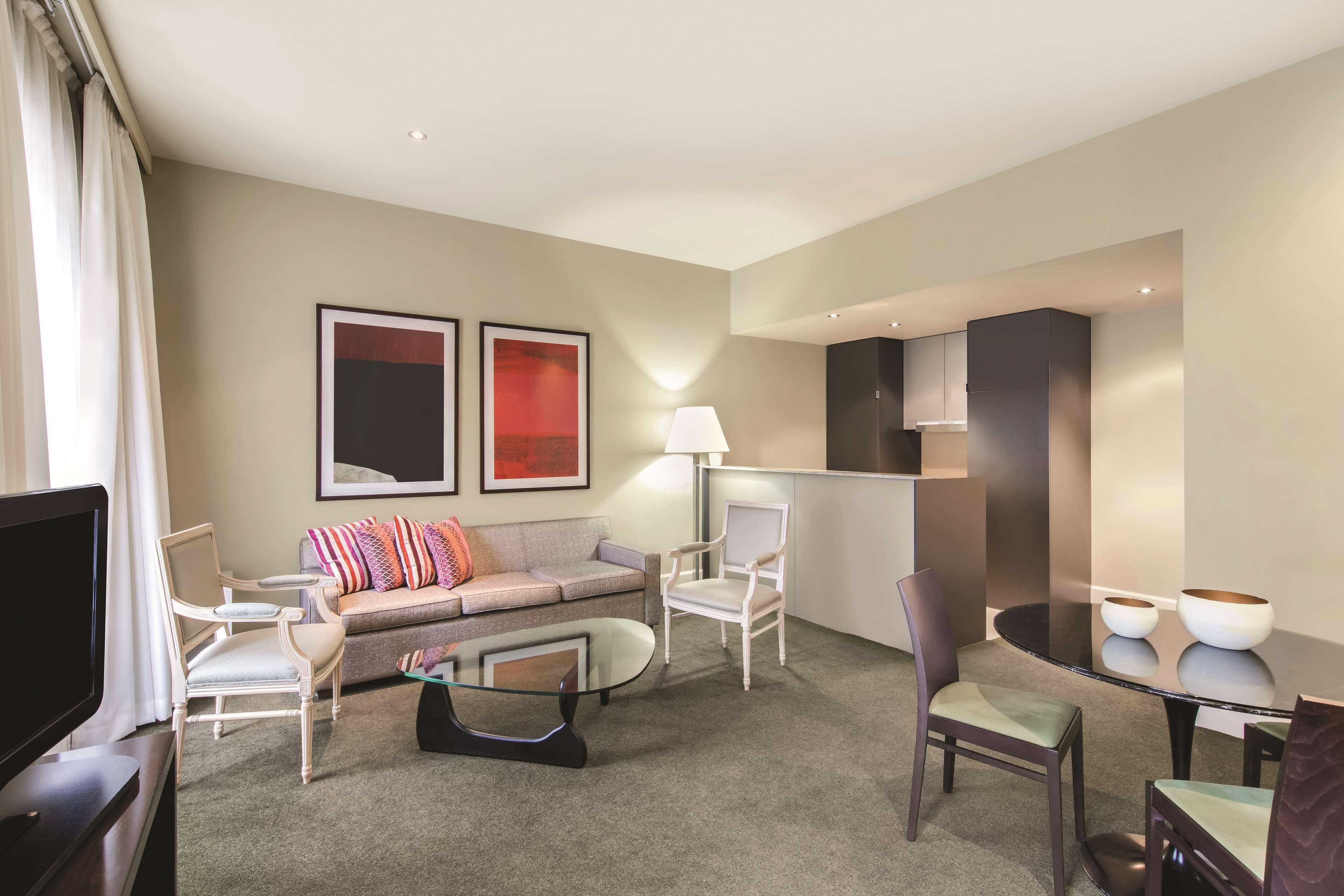 A modern hotel room with a sofa, armchairs, a glass coffee table, a dining table with two bowls, and a flat-screen TV.