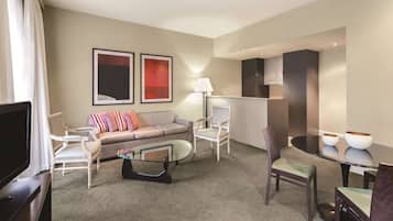 A modern hotel room with a sofa, armchairs, a glass coffee table, a dining table with two bowls, and a flat-screen TV.
