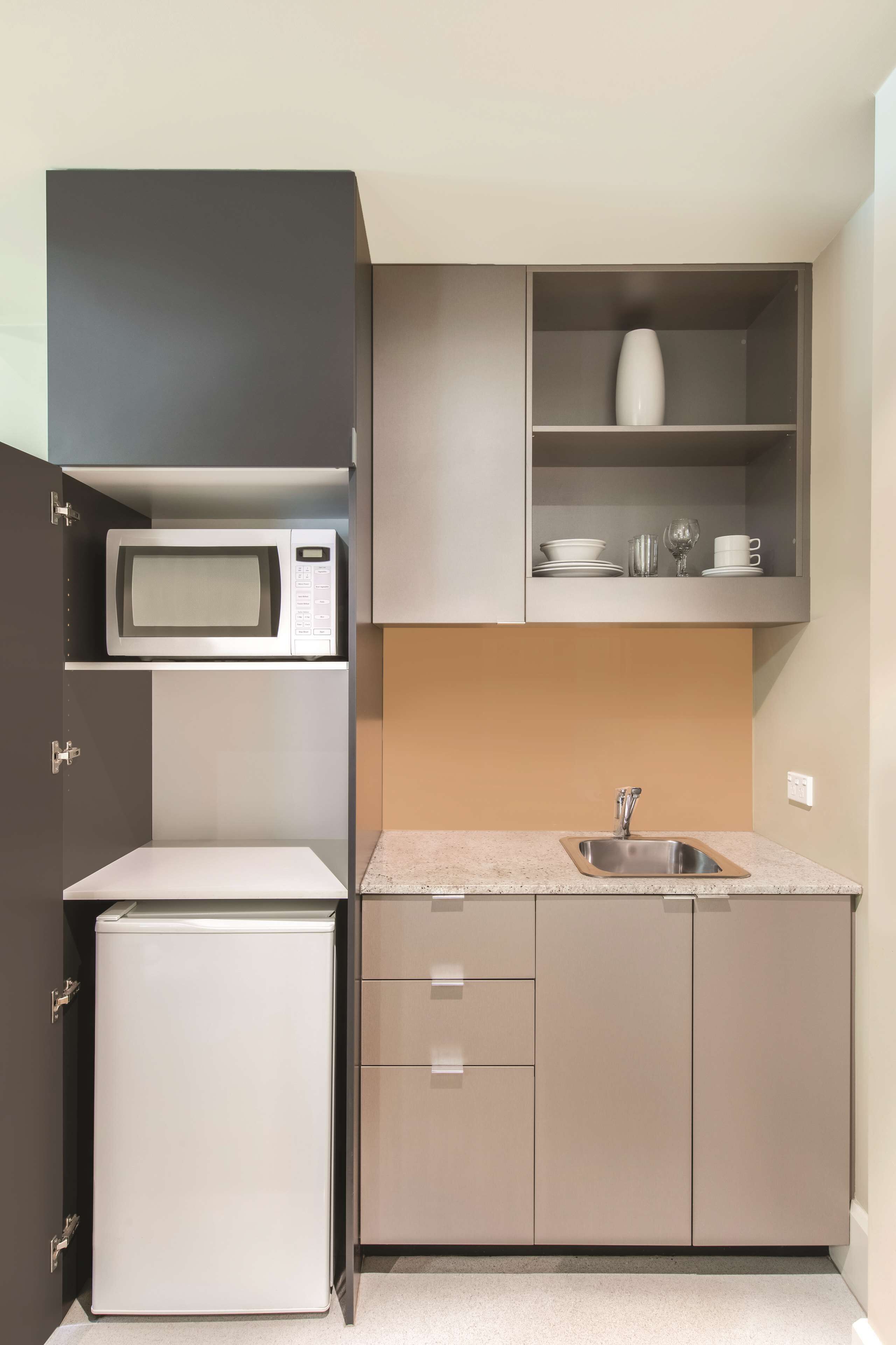 A compact kitchen with a built-in microwave, a small refrigerator, a sink, and a shelf with dishes and glassware.