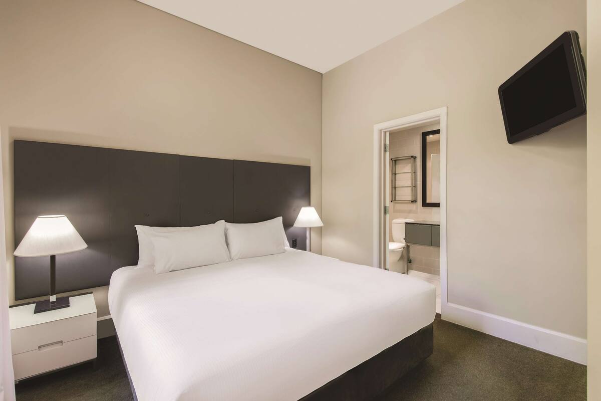 A hotel room with a large bed, two bedside tables with lamps, a flat-screen TV mounted on the wall, and a bathroom visible through an open door.