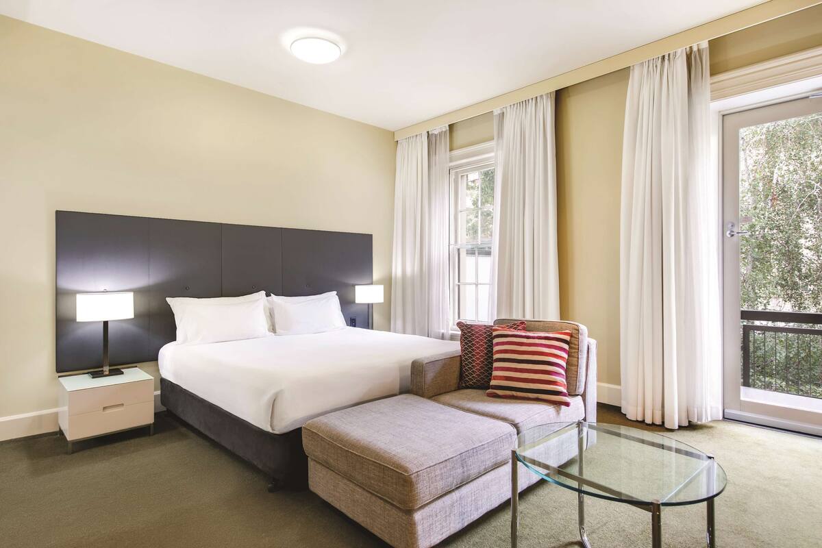 A hotel room with a large bed, a glass coffee table, a sofa, and a nightstand with a lamp.