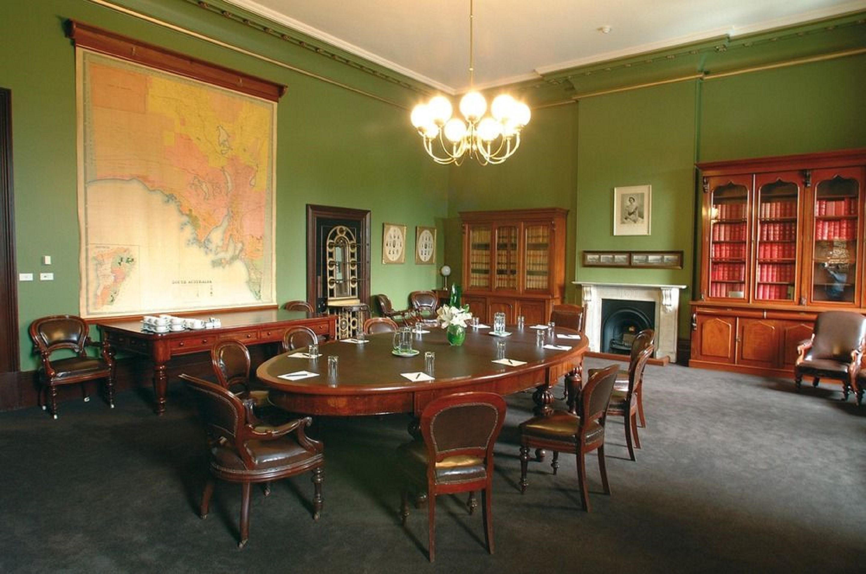 A formal meeting room with a large oval table, wooden chairs, a map on the wall, a fireplace, and bookshelves.