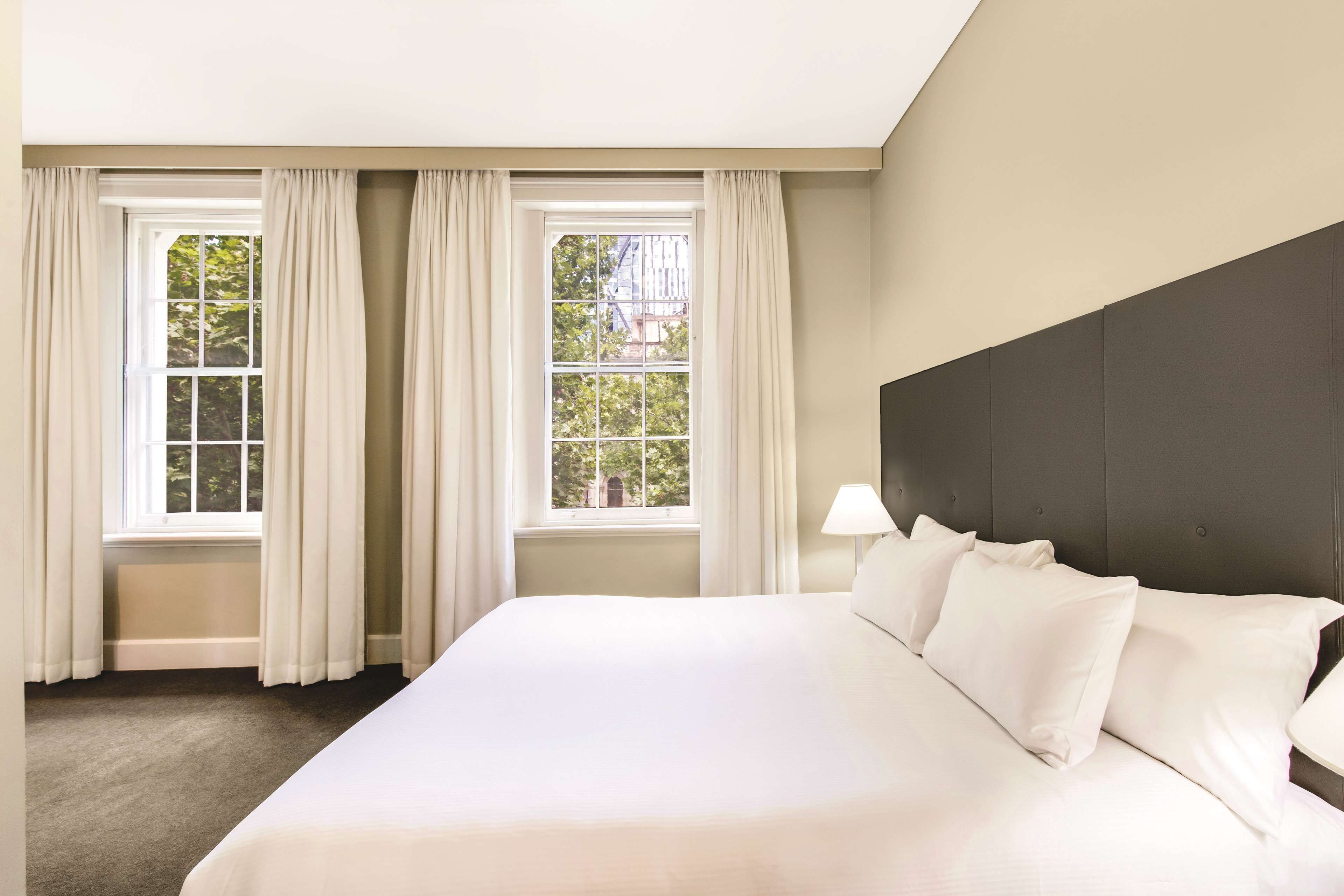 A hotel room with a large bed, two windows with curtains, a dark headboard, and a lamp on the nightstand.