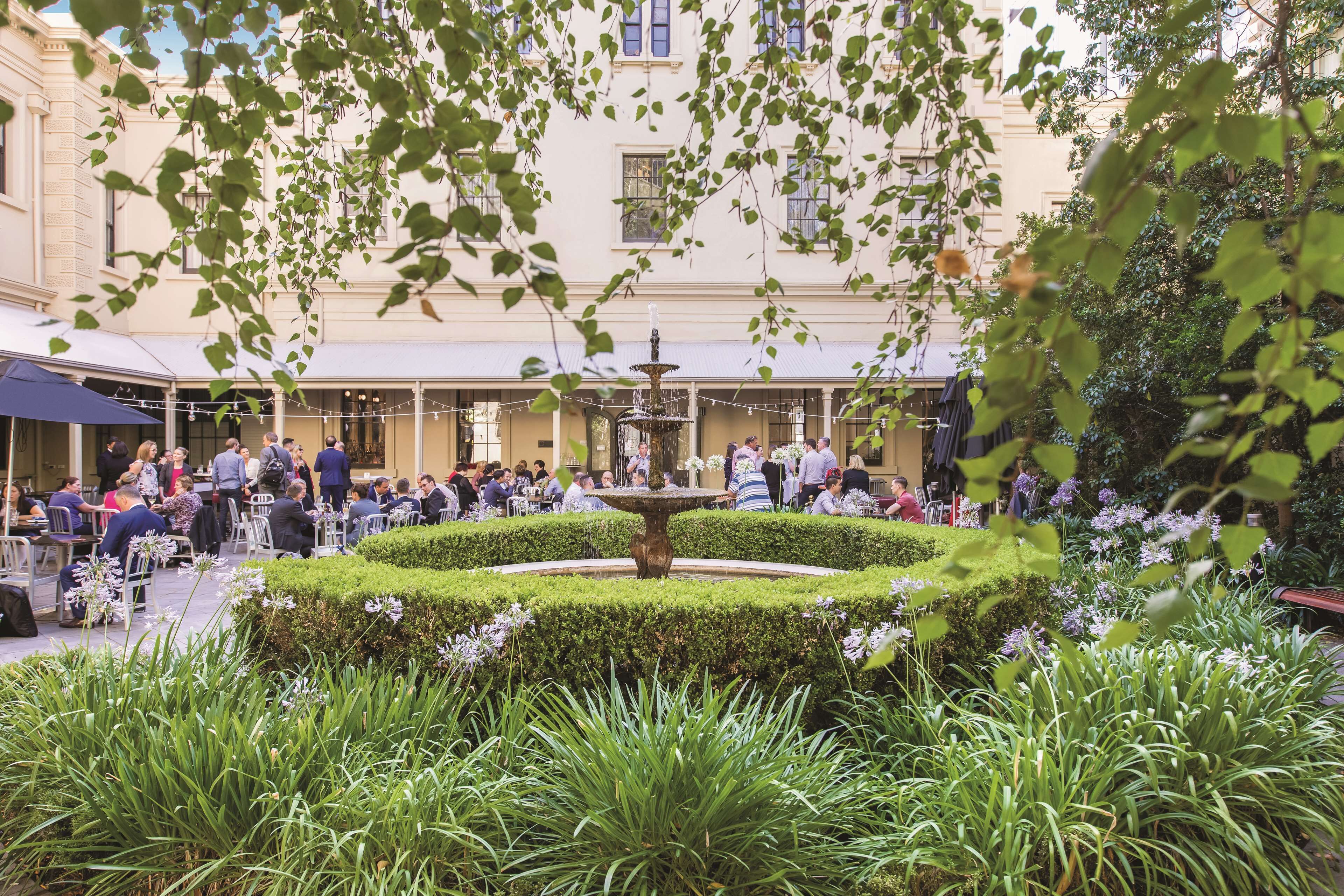 An outdoor café with a central fountain, surrounded by neatly trimmed hedges and lush greenery.