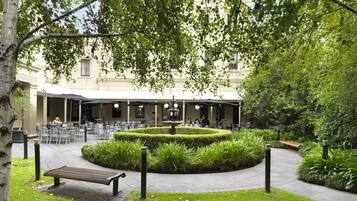 An outdoor cafe area with tables and chairs, a central fountain, and a circular hedge.